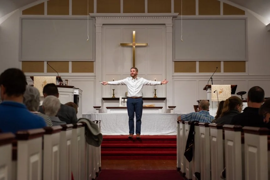 A man standing at an altar with arms outstretched inside a church, with an audience seated in pews watching.