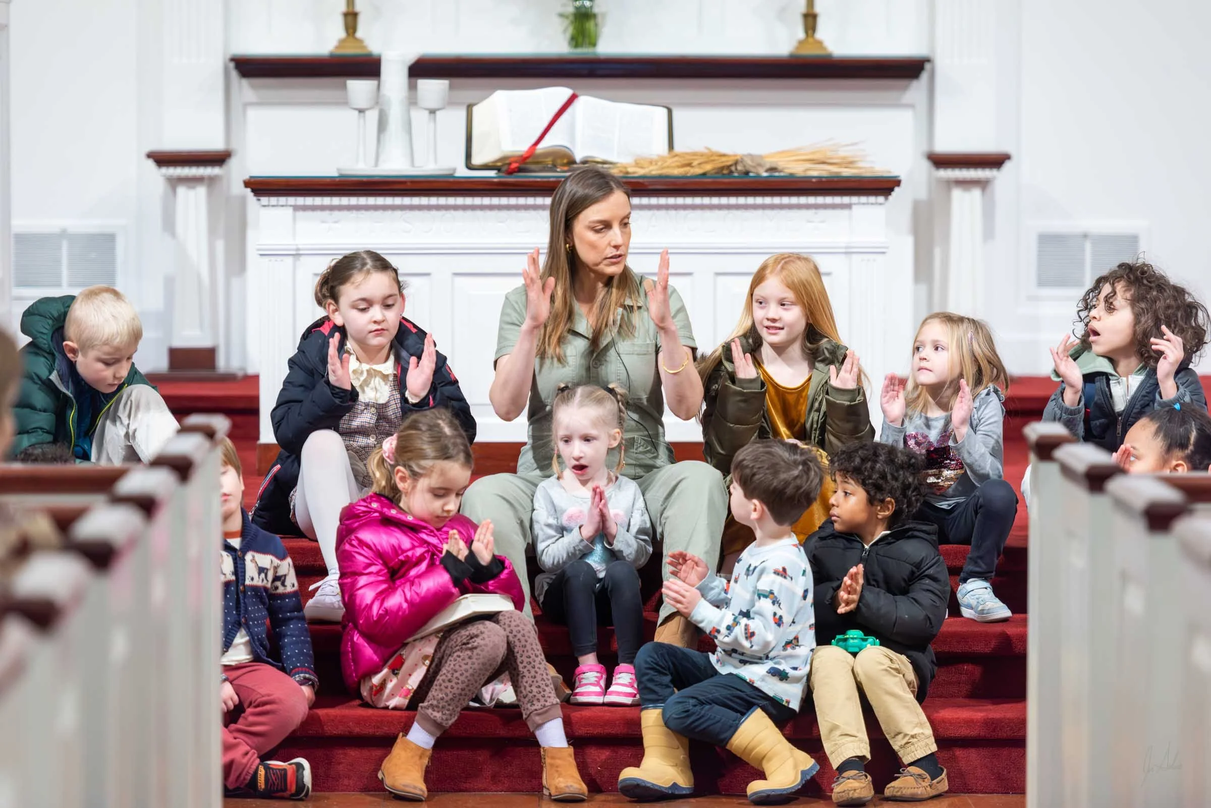 A woman sits on red-carpeted stairs surrounded by children, with her hands raised and eyes closed, possibly leading a prayer or meditation inside a church or similar setting, with a decorated fireplace and religious artifacts behind.