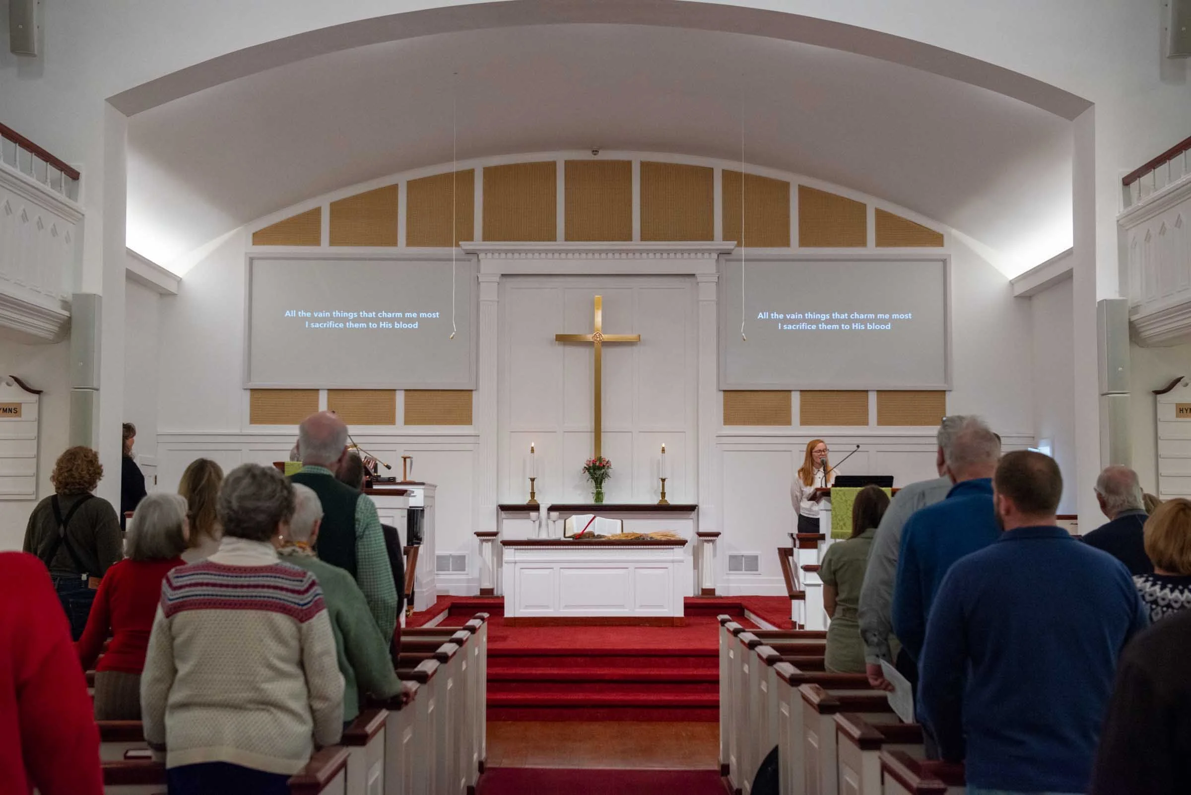 Interior of a church with a congregation listening to a woman singing at a microphone. The altar has a cross, candles, and flowers. Projected lyrics appear on screens on either side of the cross.