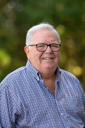 A smiling man with white hair and glasses, wearing a blue checkered shirt, standing outdoors with blurred green foliage in the background.