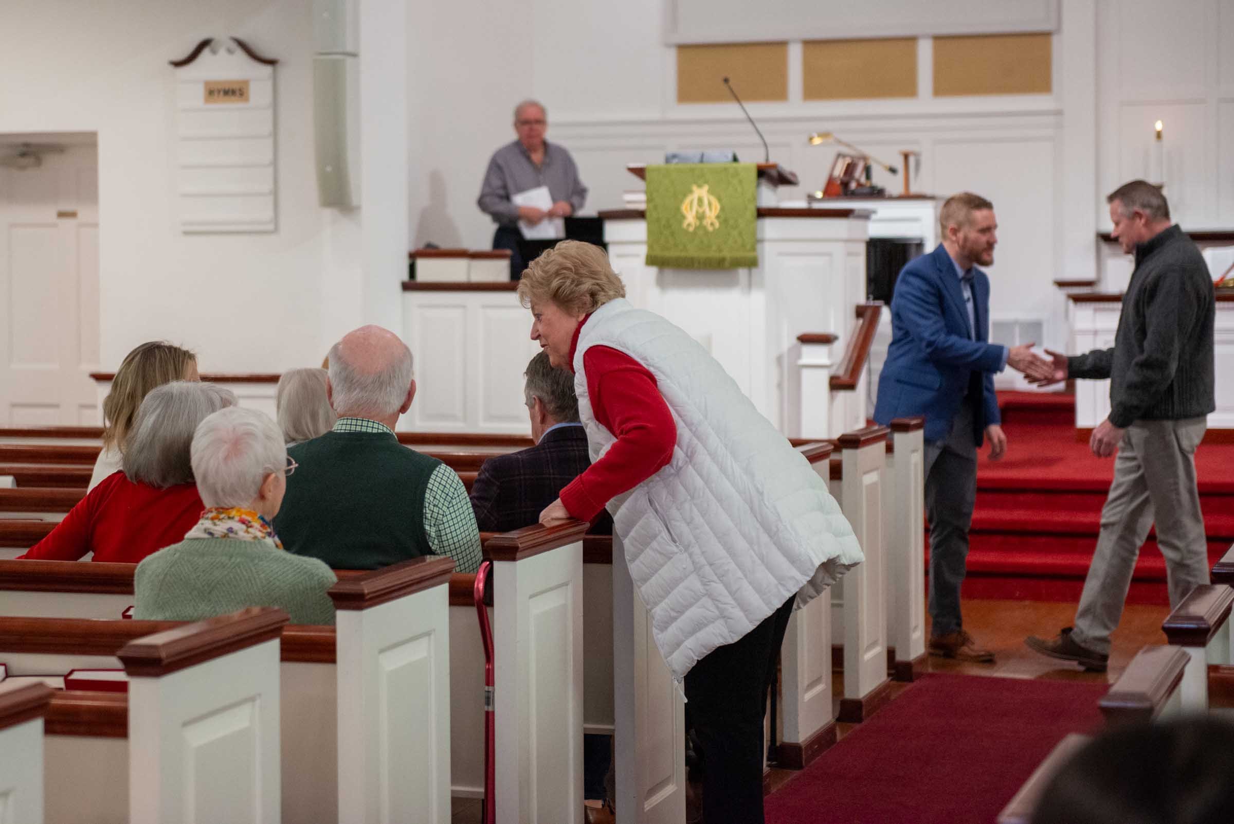 People gathered inside a church, with some seated in pews and others shaking hands. A man is standing behind the pulpit, which is decorated with a green cloth with a gold emblem.
