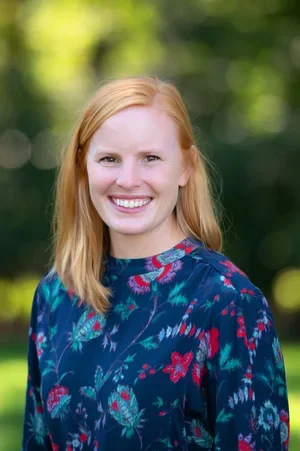 A woman with shoulder-length red hair smiling outdoors, wearing a dark blue floral blouse.