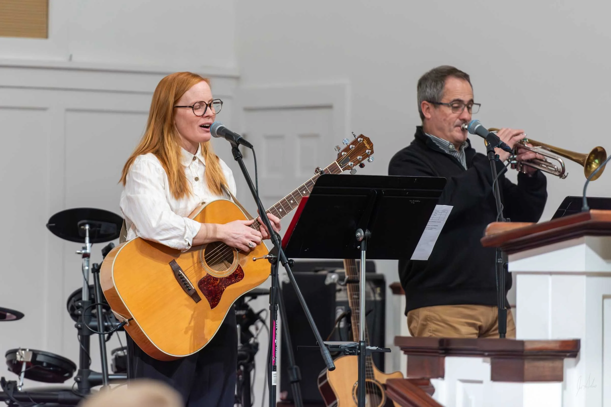 A woman with red hair and glasses playing an acoustic guitar and singing into a microphone. Next to her is a man with glasses playing a trumpet. They are performing in a white-walled room with music stands and musical equipment.
