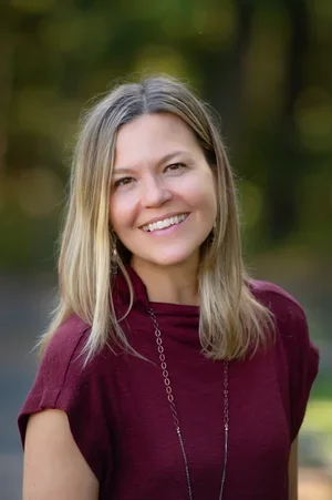 A woman with shoulder-length blonde hair and a maroon top standing outdoors with trees in the background, smiling at the camera.