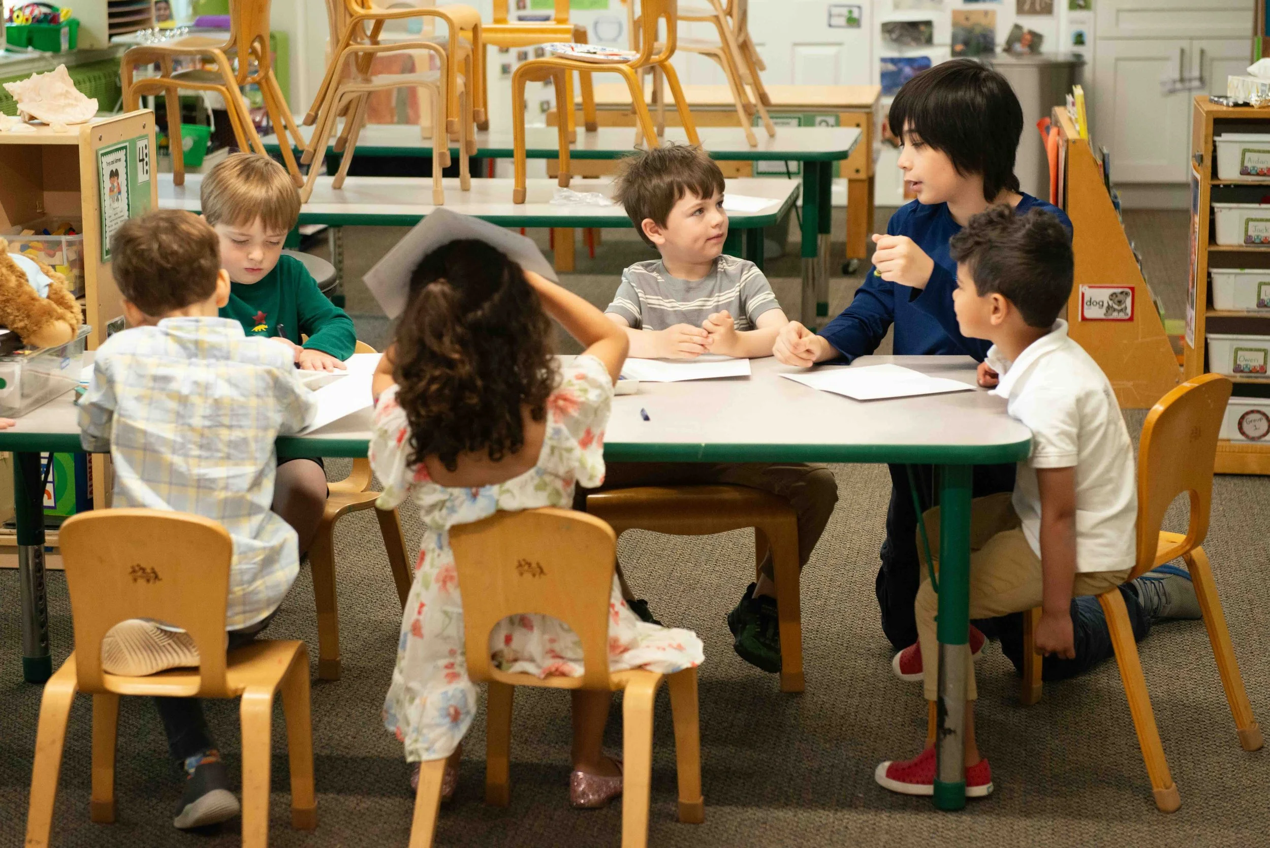 Young children sitting at a table in a classroom, engaging with a teacher.