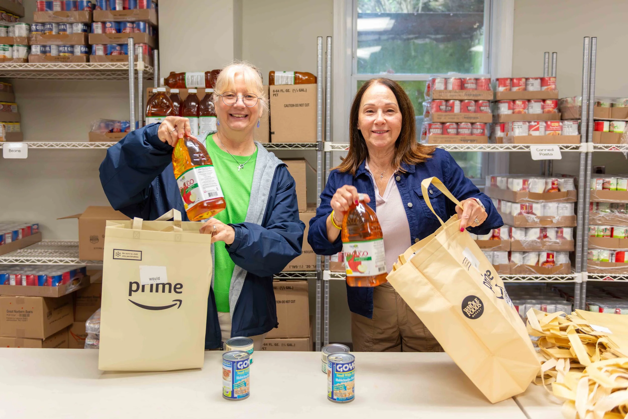 Two women standing behind a white table in a food pantry, smiling while holding canned goods and paper bags, with shelves of canned and bottled food in the background.