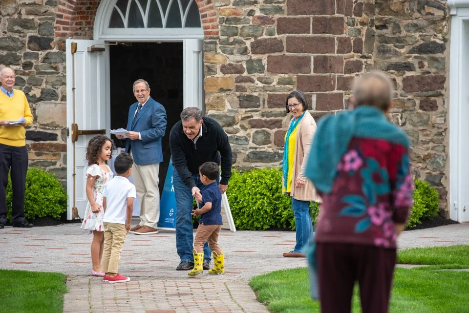 Group of children and adults outside a stone building, with a man handing something to a young boy, others smiling and engaging in conversation.