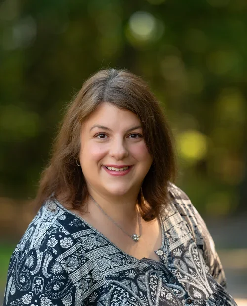 Portrait of a smiling woman with shoulder-length brown hair, wearing a patterned blouse, standing outdoors with a blurred green background.