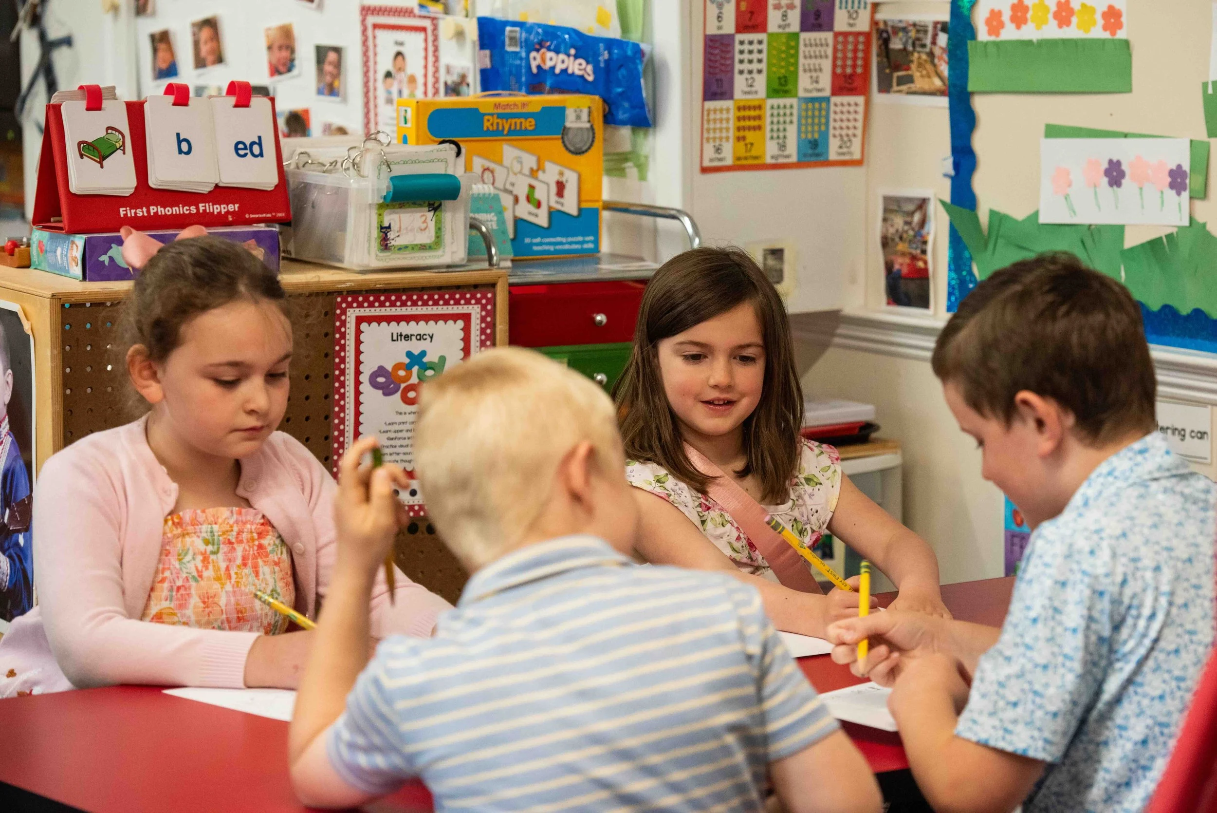 Children sitting around a table in a classroom, engaged in a learning activity. The classroom walls are decorated with educational posters, children's photographs, and colorful artwork. There are educational materials on the table and shelves in the background.