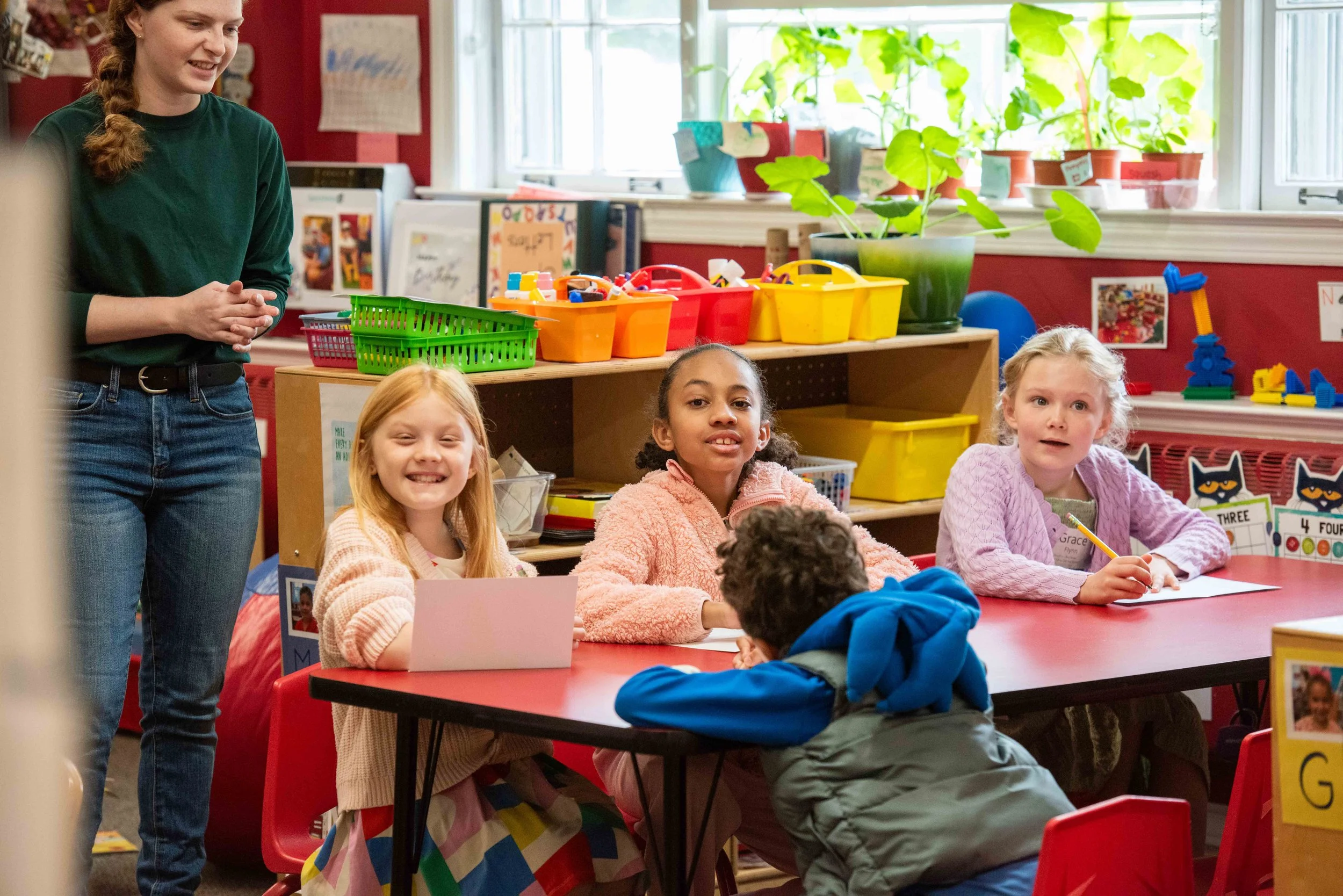 Children sitting at a classroom table during a lesson, with a teacher standing nearby, colorful storage bins, plants on the windowsill, and classroom decorations.