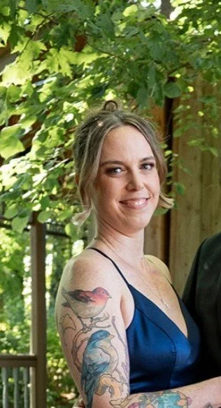 A woman with tattoos on her left arm, wearing a blue dress, standing outdoors with green foliage in the background.