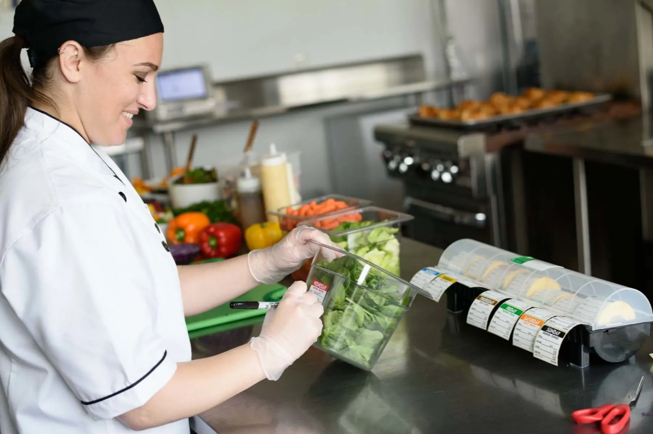 A smiling woman in a chef's coat and gloves prepares a salad in a commercial kitchen, with various fresh vegetables and food containers on the counter.