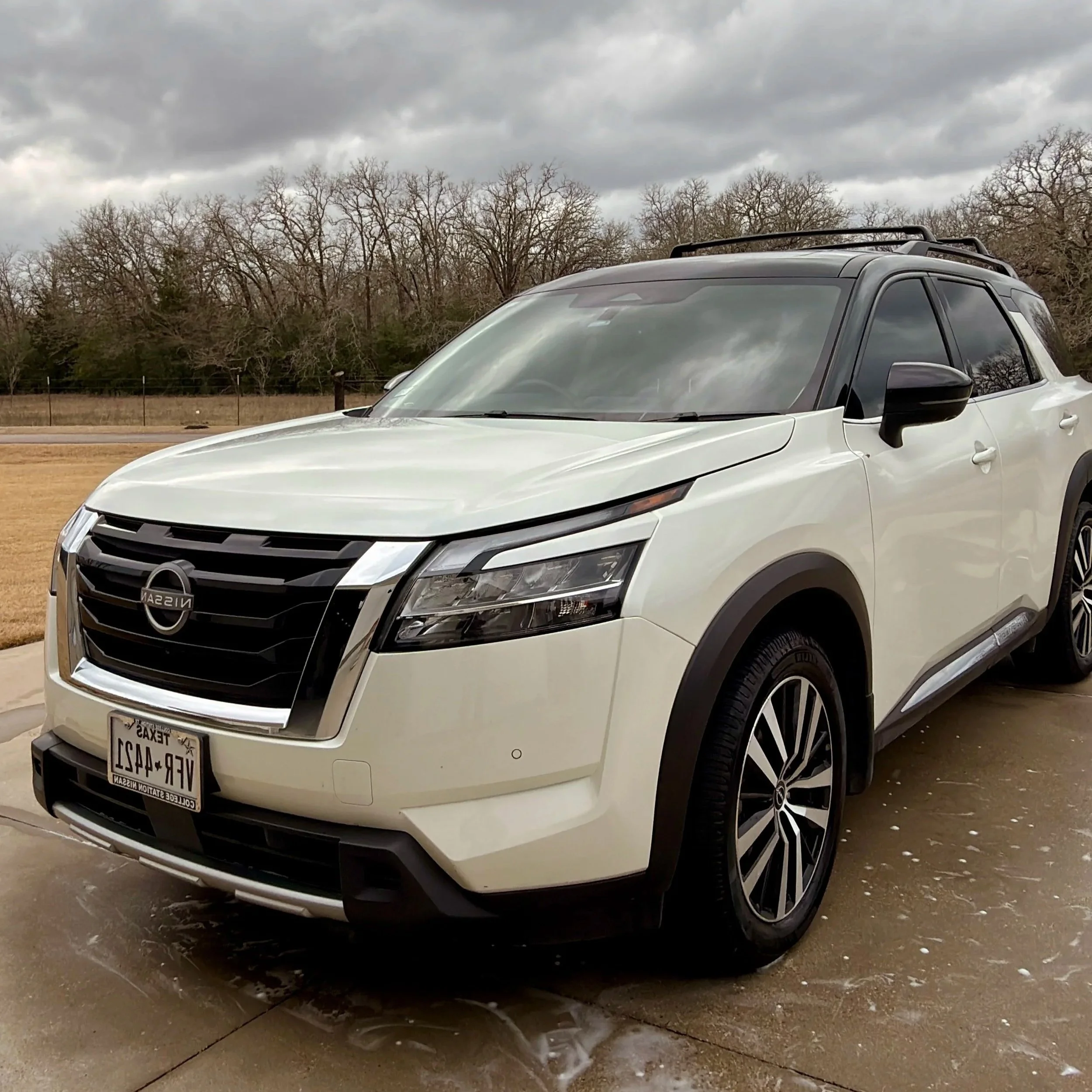 White Nissan SUV parked outdoors on a wet pavement with cloudy sky and leafless trees in the background.