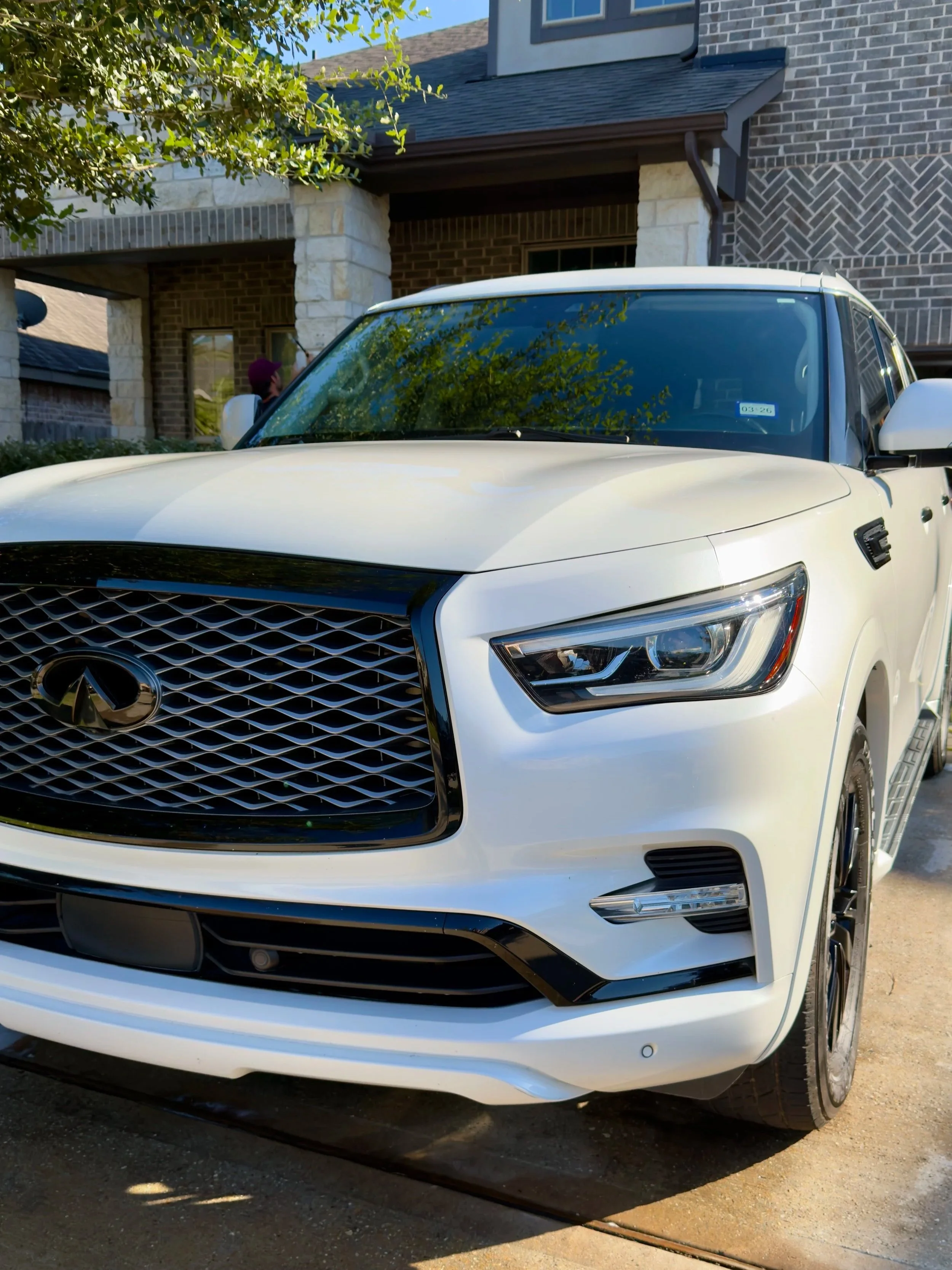 Front view of a white Infiniti SUV parked in front of a brick house with trees and plants nearby.