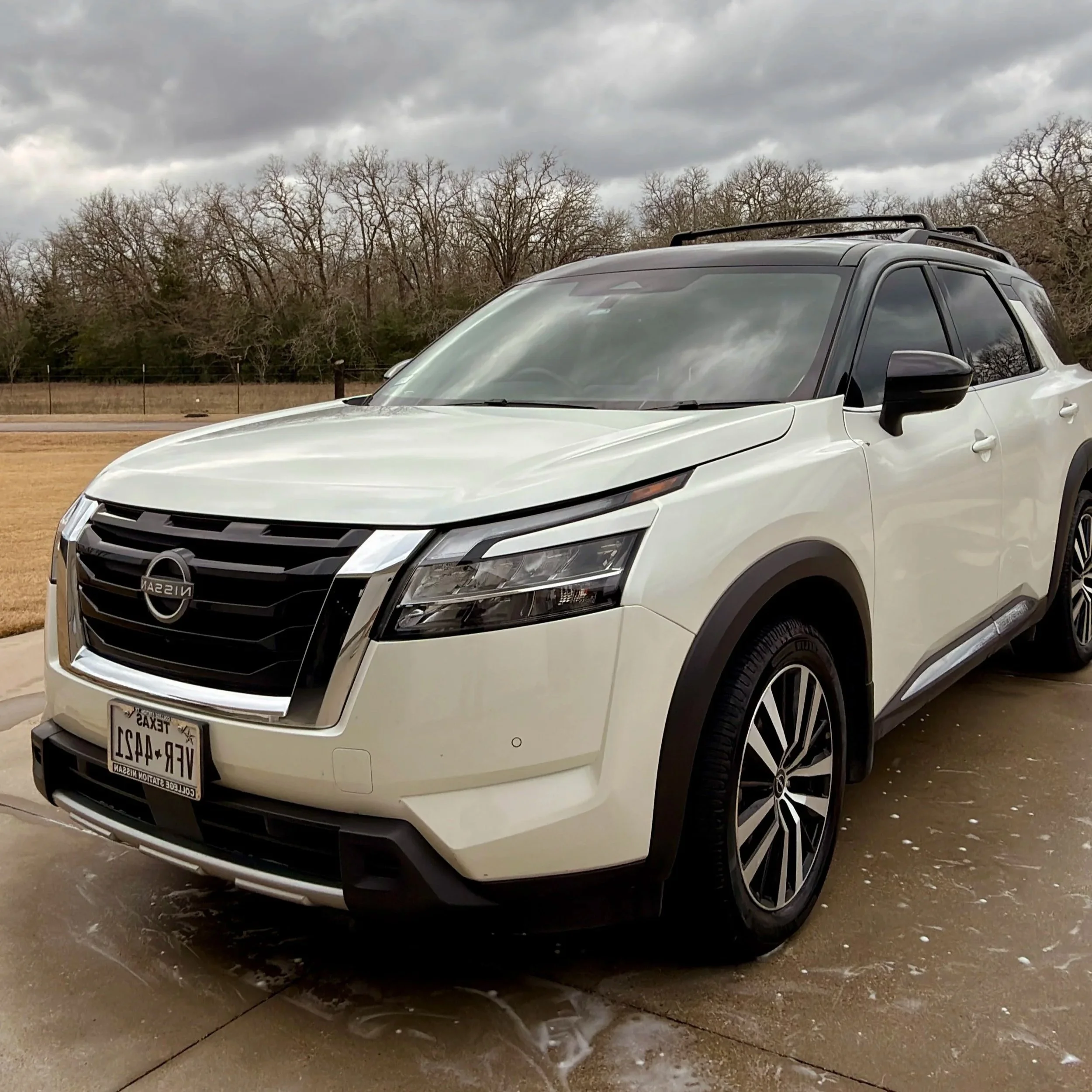 A white Nissan SUV parked outdoors on a concrete surface with cloudy sky and leafless trees in the background.