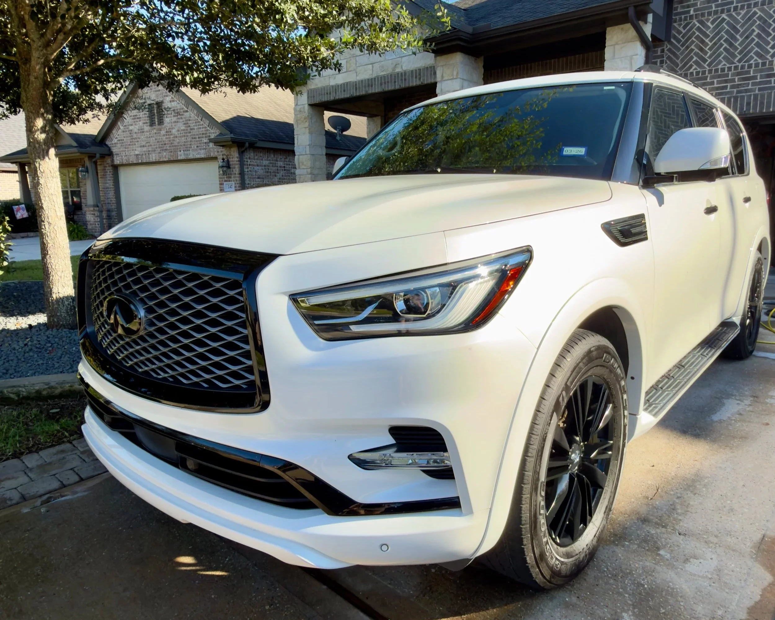 A white luxury SUV parked in front of a house with brick and stone exterior, green trees, and a driveway.