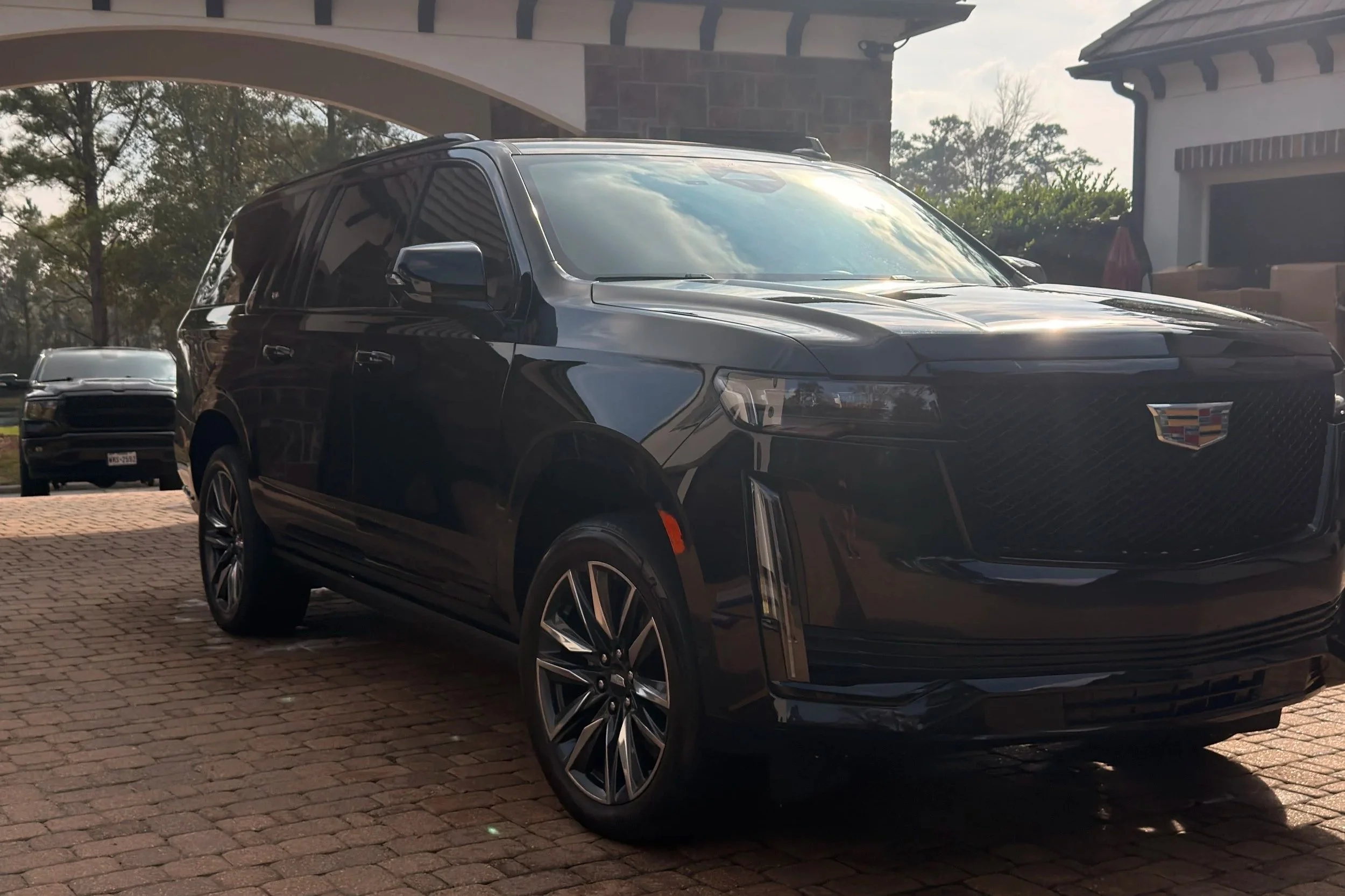 Black Cadillac SUV parked on a brick driveway with other cars and a building in the background.