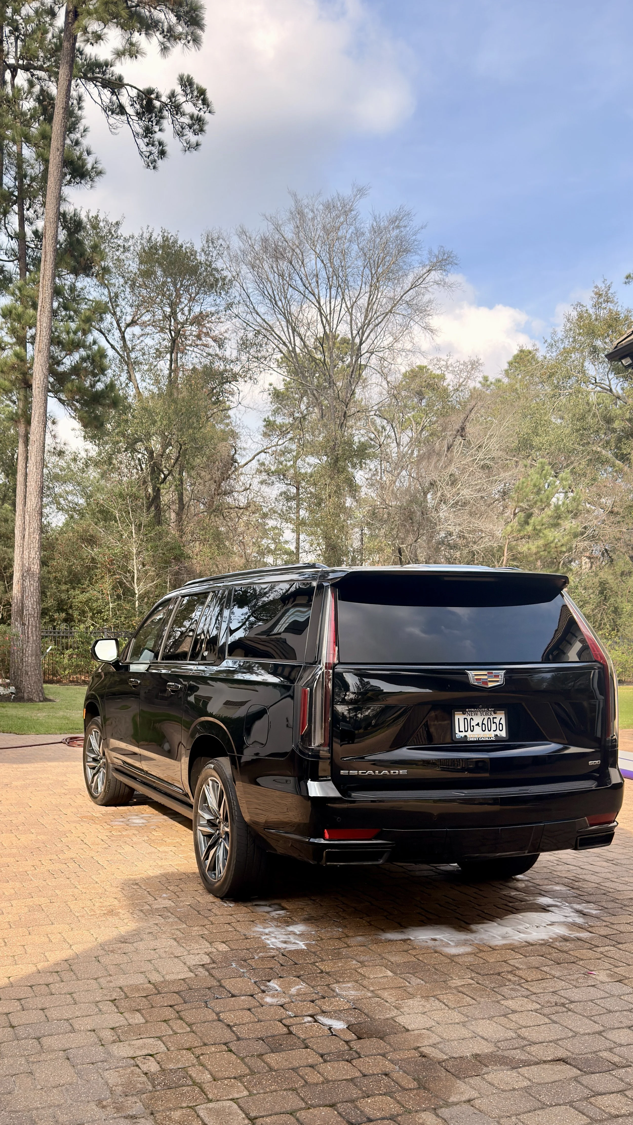 A black Cadillac Escalade parked on a brick driveway with some water on the ground, surrounded by trees and a partly cloudy sky.