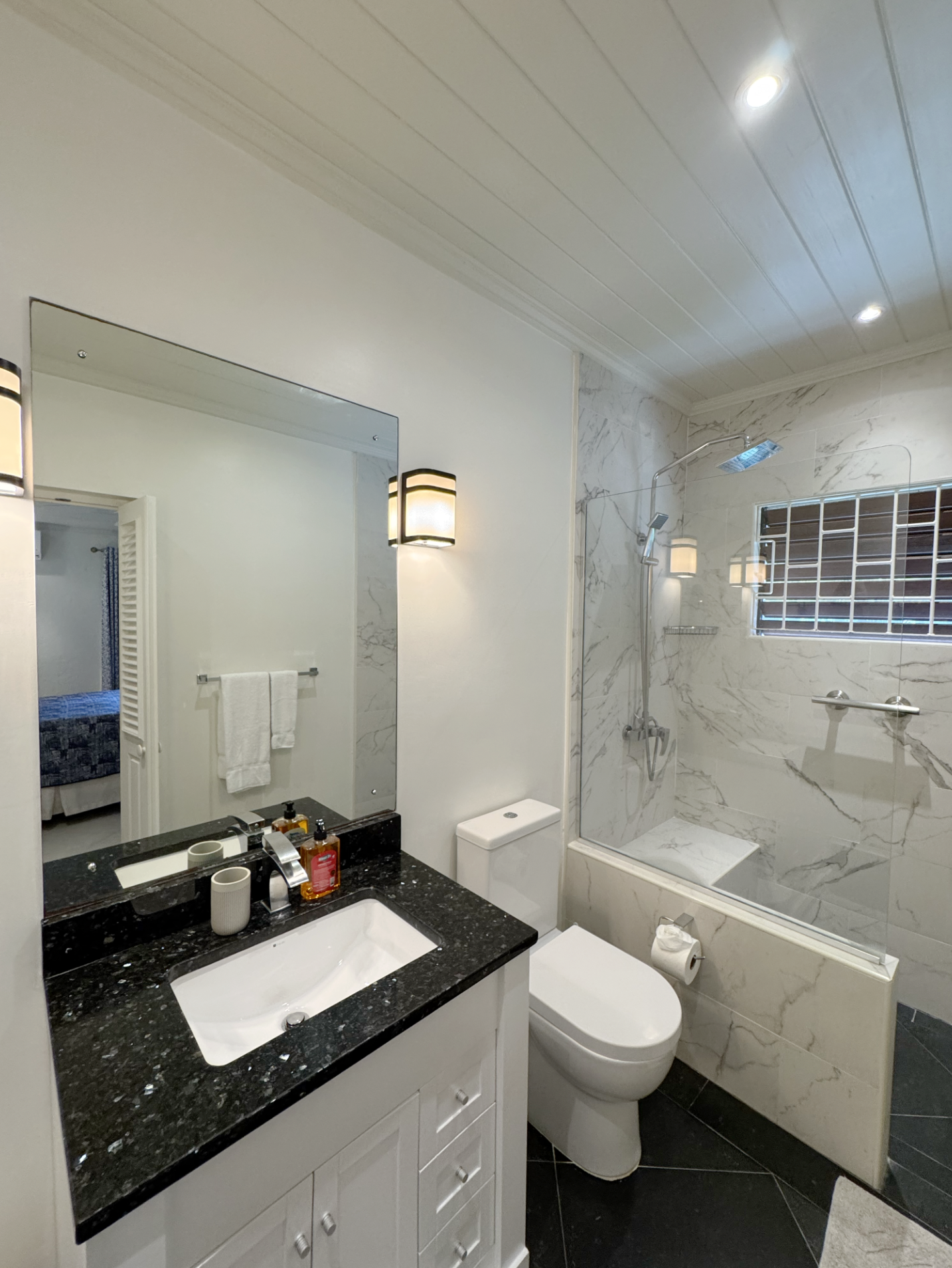 Bathroom with a black granite countertop sink, white cabinetry, a toilet, a bathtub with marble walls, a glass shower partition, a window with metal bars, and wall-mounted light fixtures.