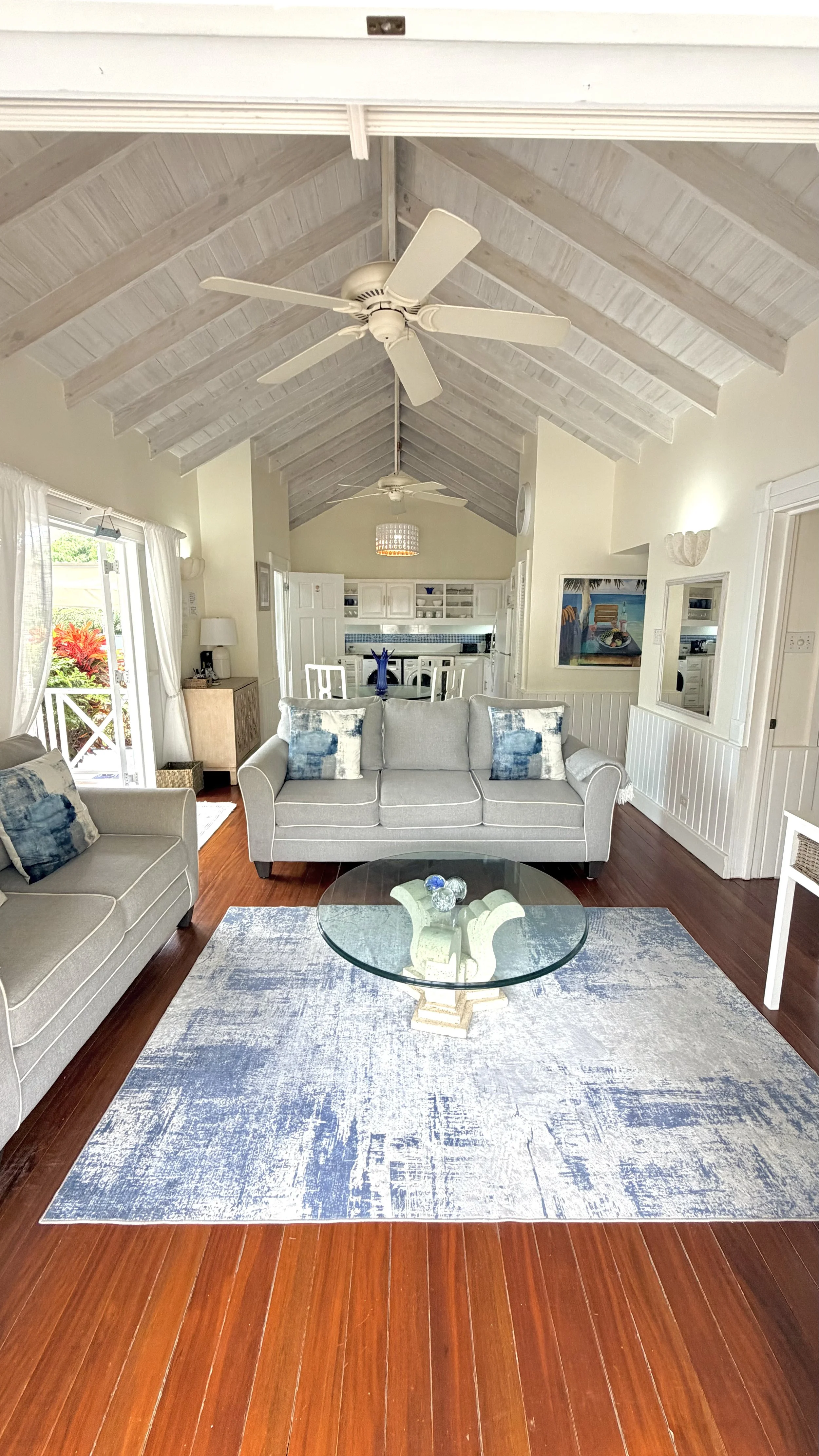 Living room with two gray sofas, blue and white pillows, a glass coffee table, a blue and white rug, and a view of the kitchen and outdoors through large windows and a sliding door. The ceiling has white beams and two ceiling fans.