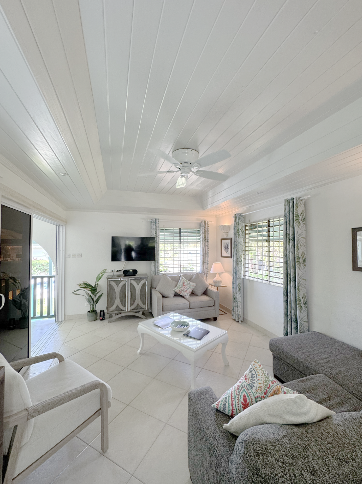 Living room with beige and gray furniture, white coffee table, TV on the wall, windows with tropical curtains, ceiling fan, and tiled floor.