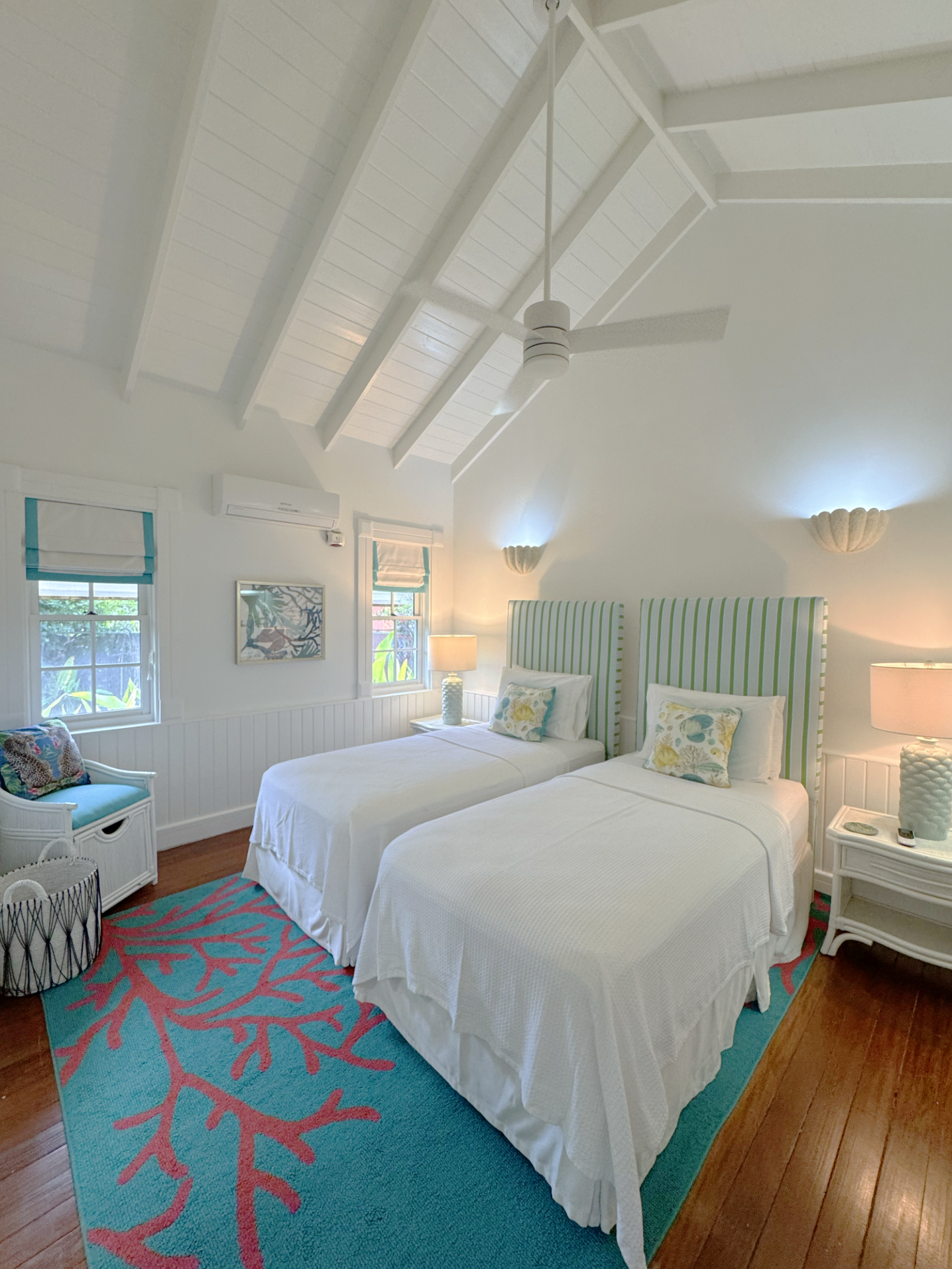A bedroom with two beds, each with green and white striped headboards and floral pillows. The room has white walls, two windows with blue and white shades, a ceiling fan, wall sconces, a colorful coral-patterned rug, a wicker chair with a floral cush