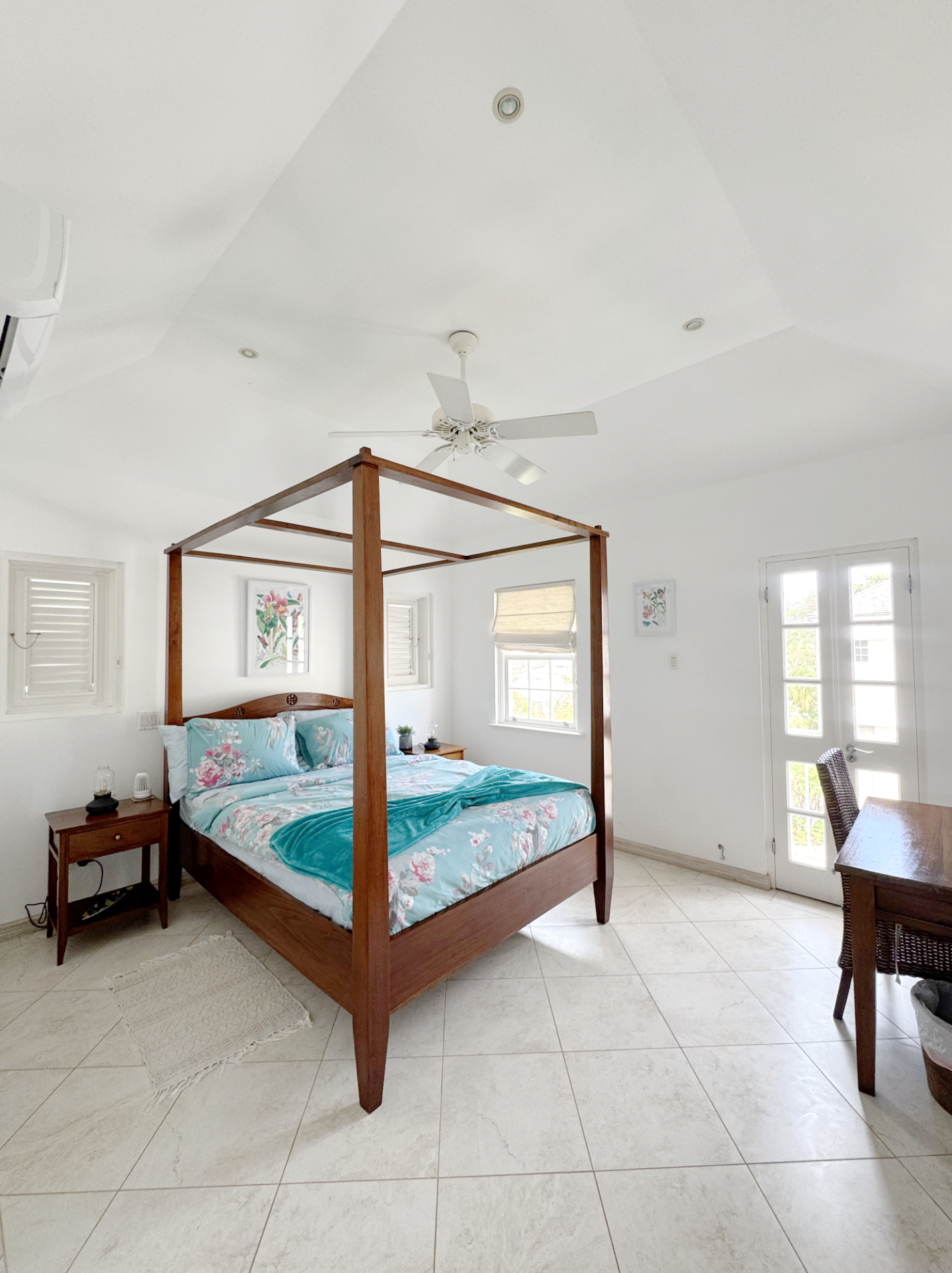 A bedroom with a wooden four-poster bed with floral bedding, a nightstand with a lamp, multiple windows, and white walls with framed artwork.