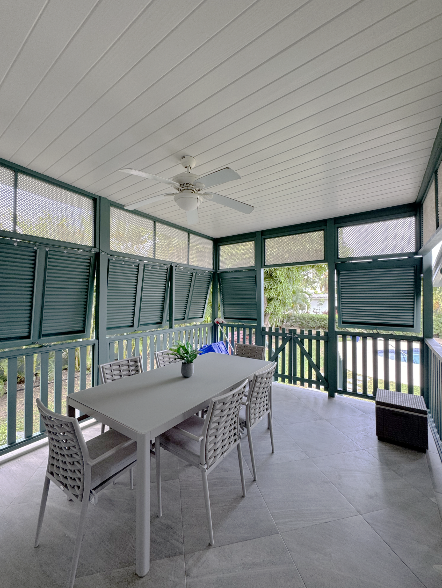 Covered porch area with a white dining table and six white chairs, green fencing with shutters, ceiling fan, potted plant on the table, and lush trees visible outside.