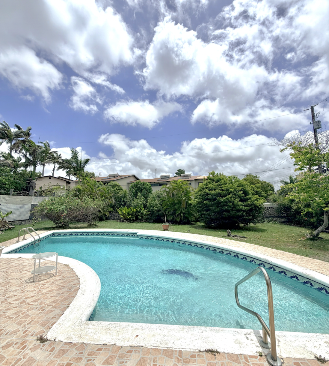 Residential backyard with a swimming pool, surrounded by greenery and trees, under a partly cloudy sky.