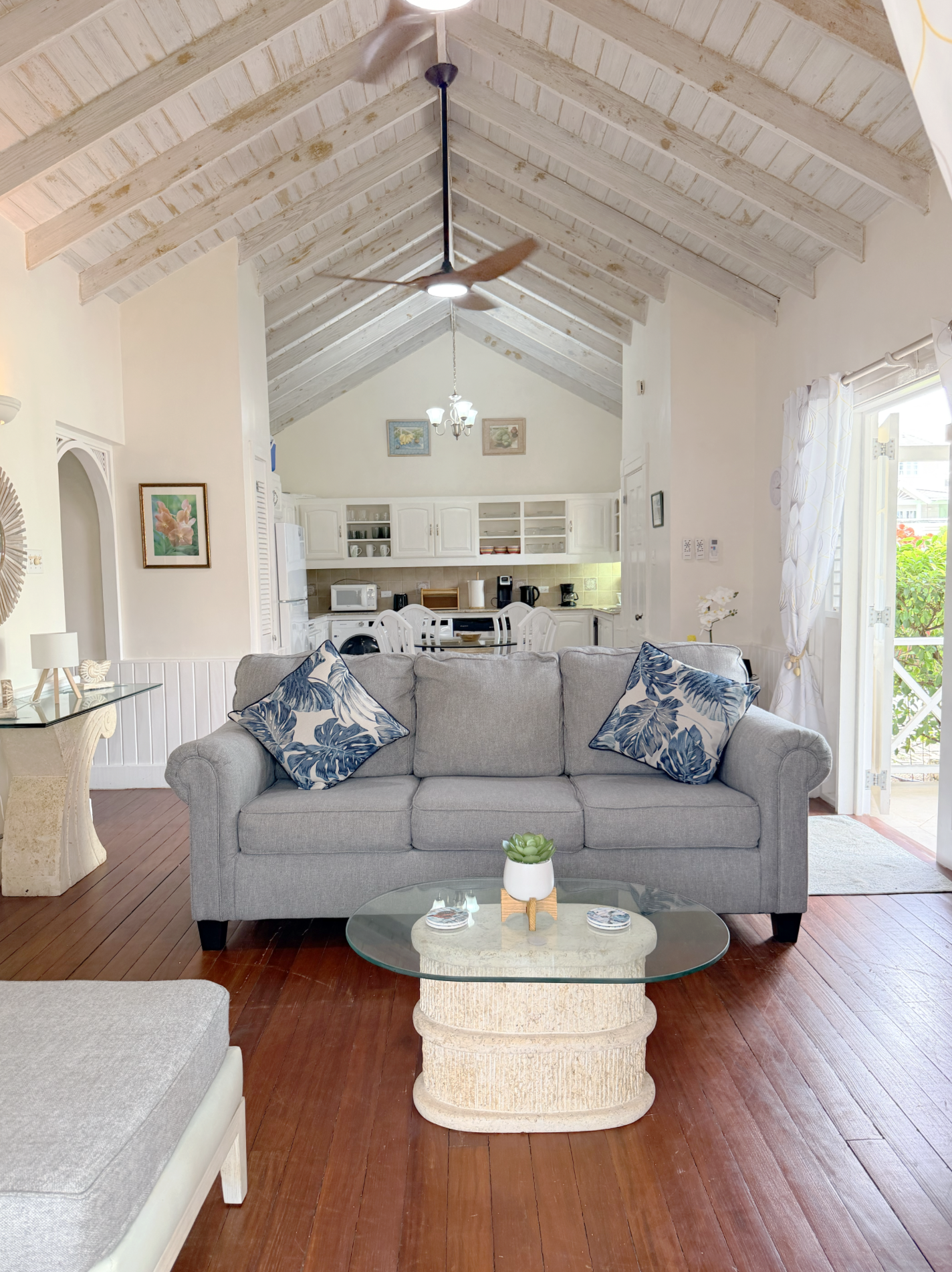 Living room with wooden floors, a gray sofa with blue and white tropical patterned pillows, glass coffee table with a small succulent plant, and a view into a kitchen with white cabinets and a dining area, bright natural light coming from a partially