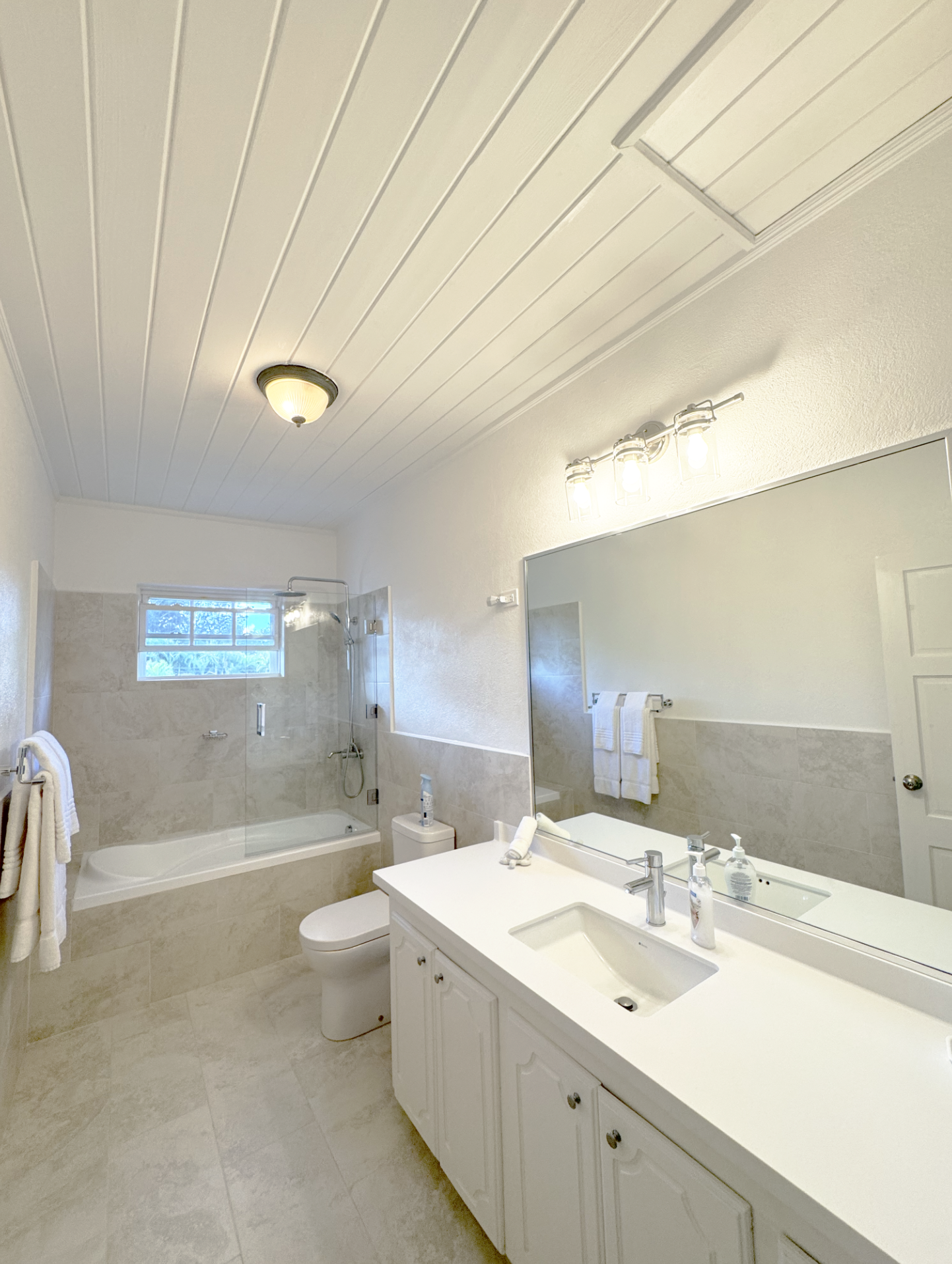 A bathroom with a white vanity, large mirror, white countertop, and sink. Behind the vanity, there is a bathtub with a glass shower door. The ceiling has white paneling, with a ceiling light fixture and modern bathroom lighting above the mirror. Towe