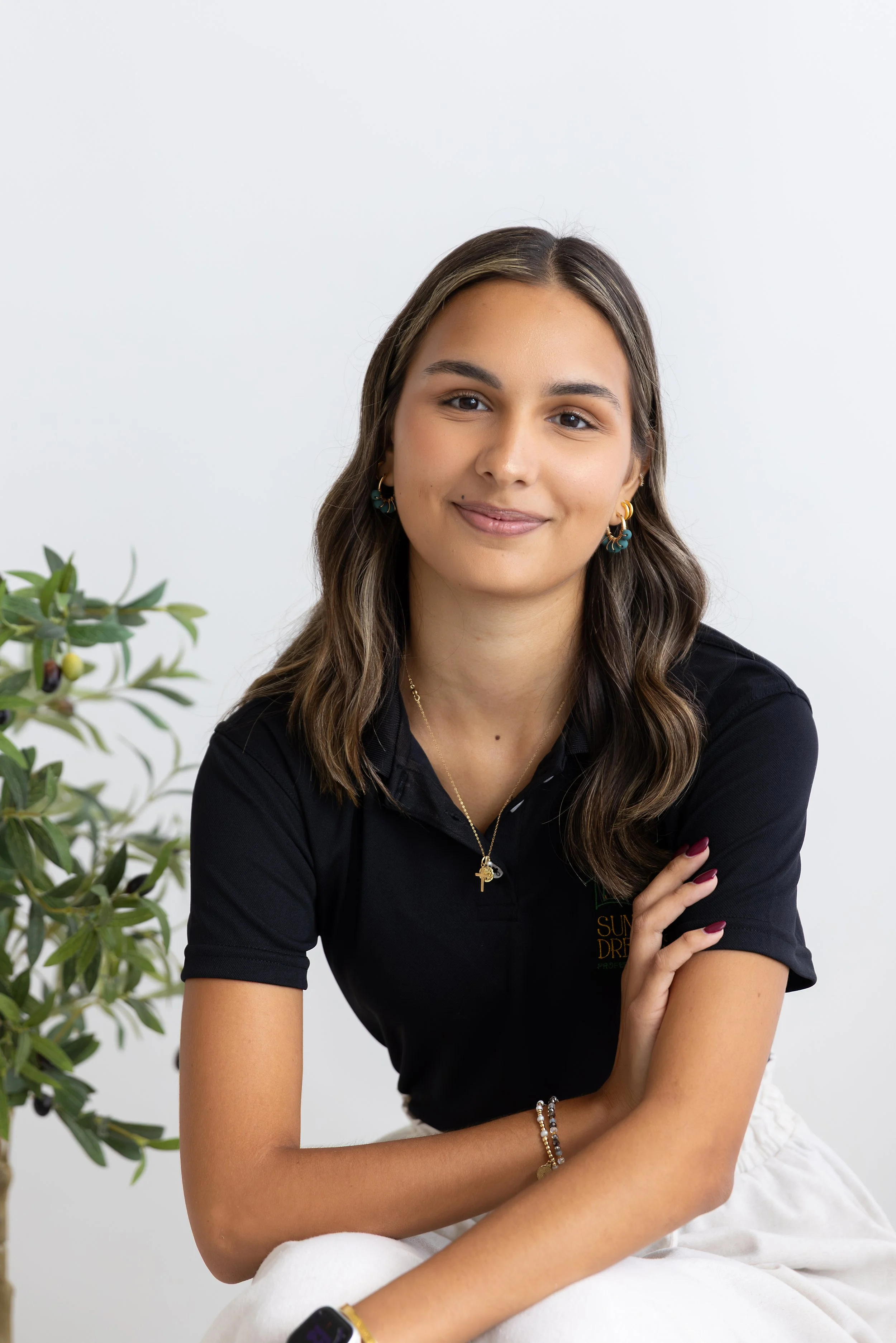 A young woman with long wavy brown hair, wearing a black polo shirt with a logo, gold earrings, and layered necklaces, sitting with her arms crossed and smiling in front of a plain white background, with a leafy plant partially visible on the left.
