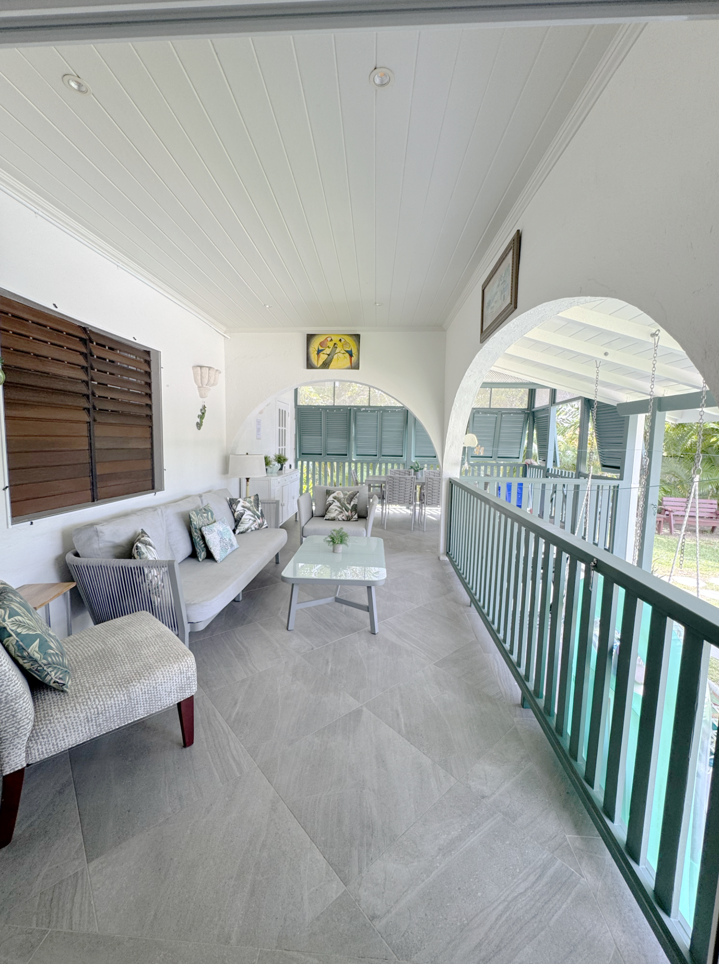 Covered balcony with white ceiling, windows with green shutters, beige and white upholstered furniture, a white coffee table, decorative pillows, and hanging swings with chains.