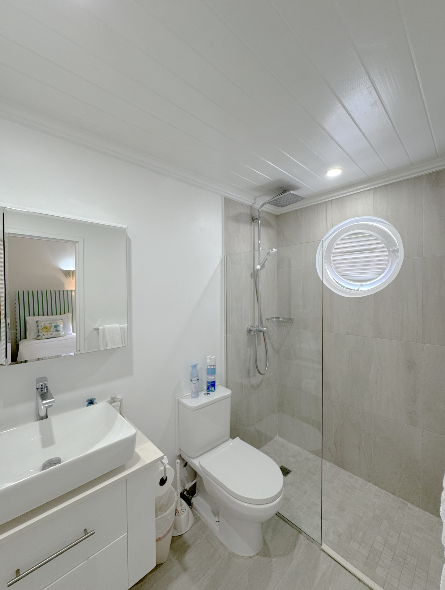 A modern bathroom with a white sink and cabinet, a toilet, a mirror, and a shower area with a window, tiled walls, and a ceiling with a light.