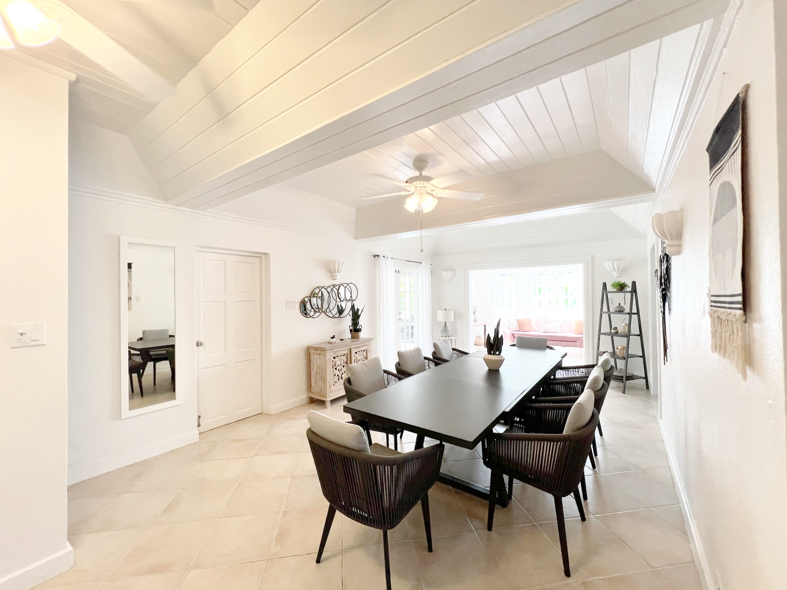 A dining room with a black dining table surrounded by nine chairs with beige cushions, a white ceiling with a fan, large windows with white curtains, and wall decor including a woven art piece and plants.