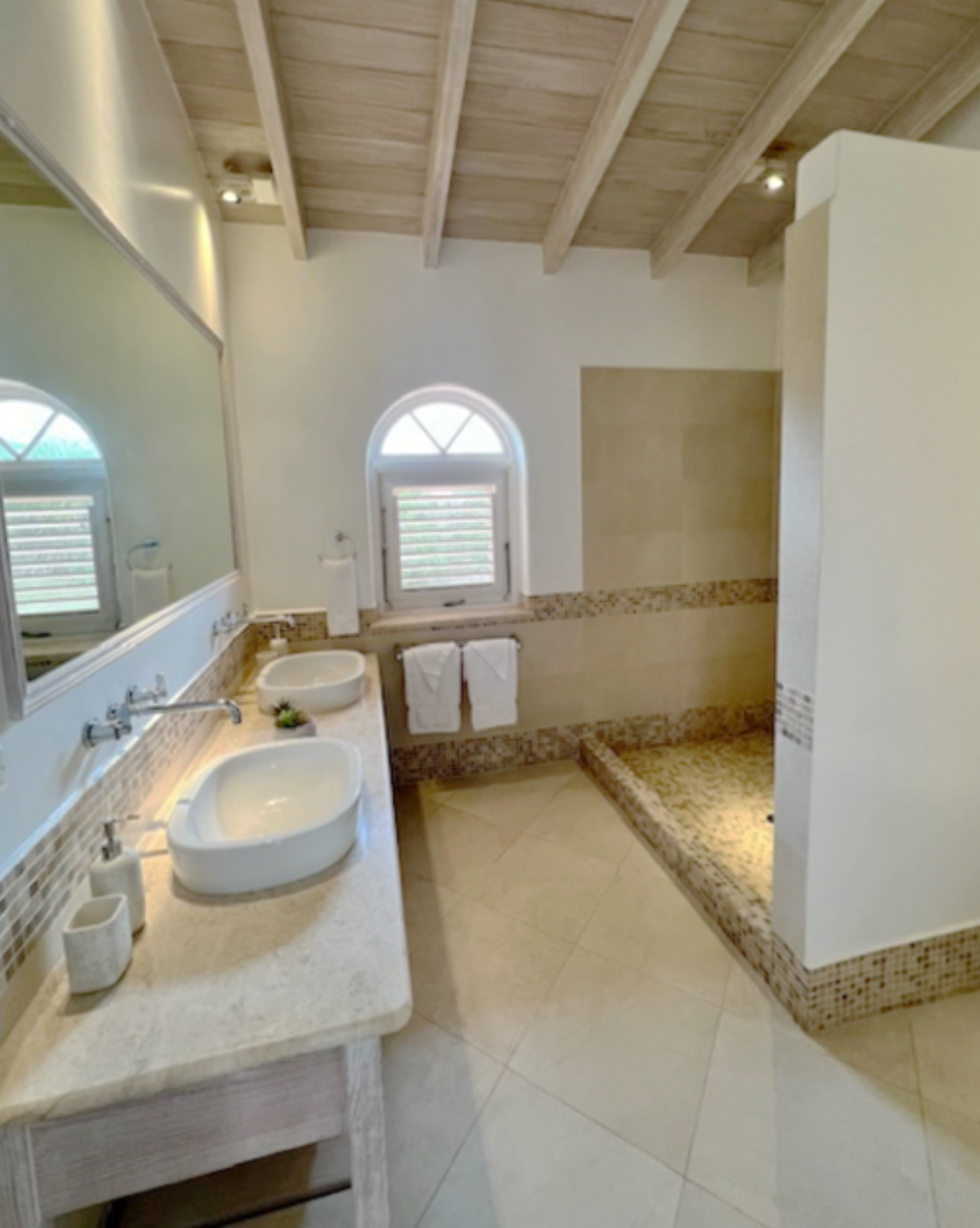 A bathroom with dual vessel sinks, a large mirror, a window with shutters, and a shower area with a bench, all featuring a neutral color scheme and wooden ceiling beams.