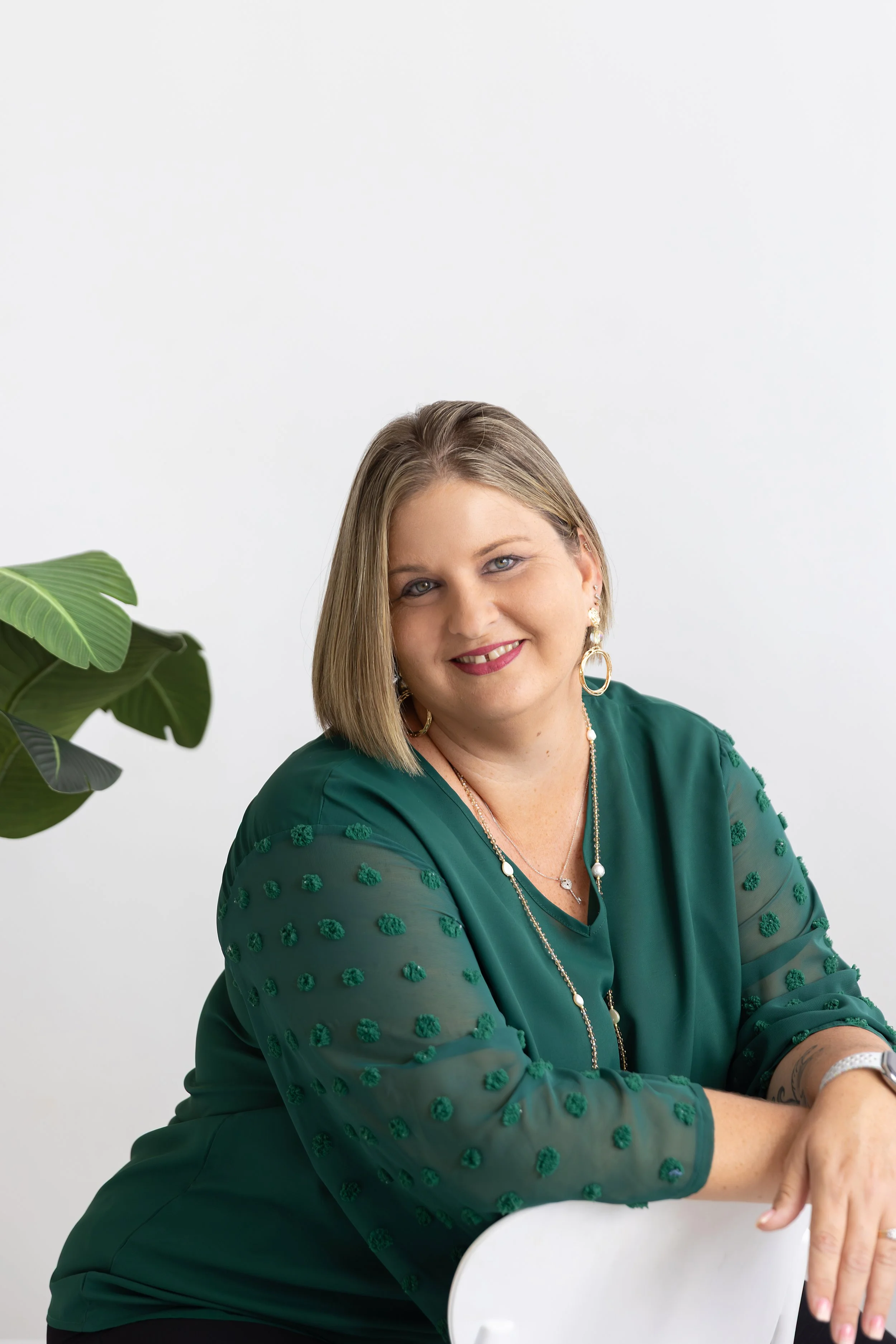 A smiling woman with shoulder-length brown hair, wearing a dark green blouse with textured polka dots, layered necklaces, and earrings, sitting with her arm resting on a white chair, against a plain white wall with a large green plant partially visible on the left.