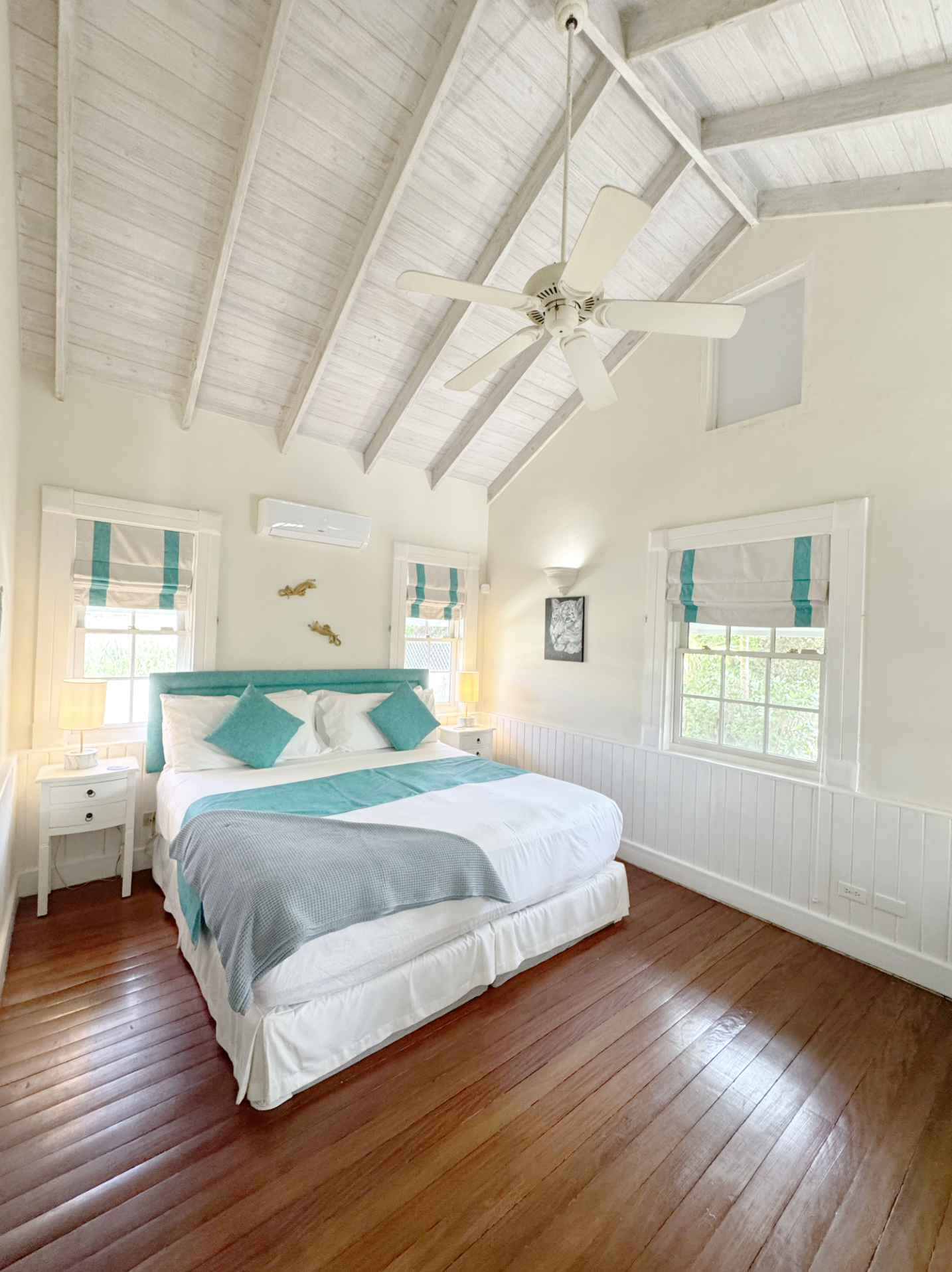 A bedroom with a vaulted white wood-paneled ceiling, a ceiling fan, two windows with striped roman shades, white walls, a bed with white bedding and turquoise accent pillows, nightstands with lamps, and hardwood floors.