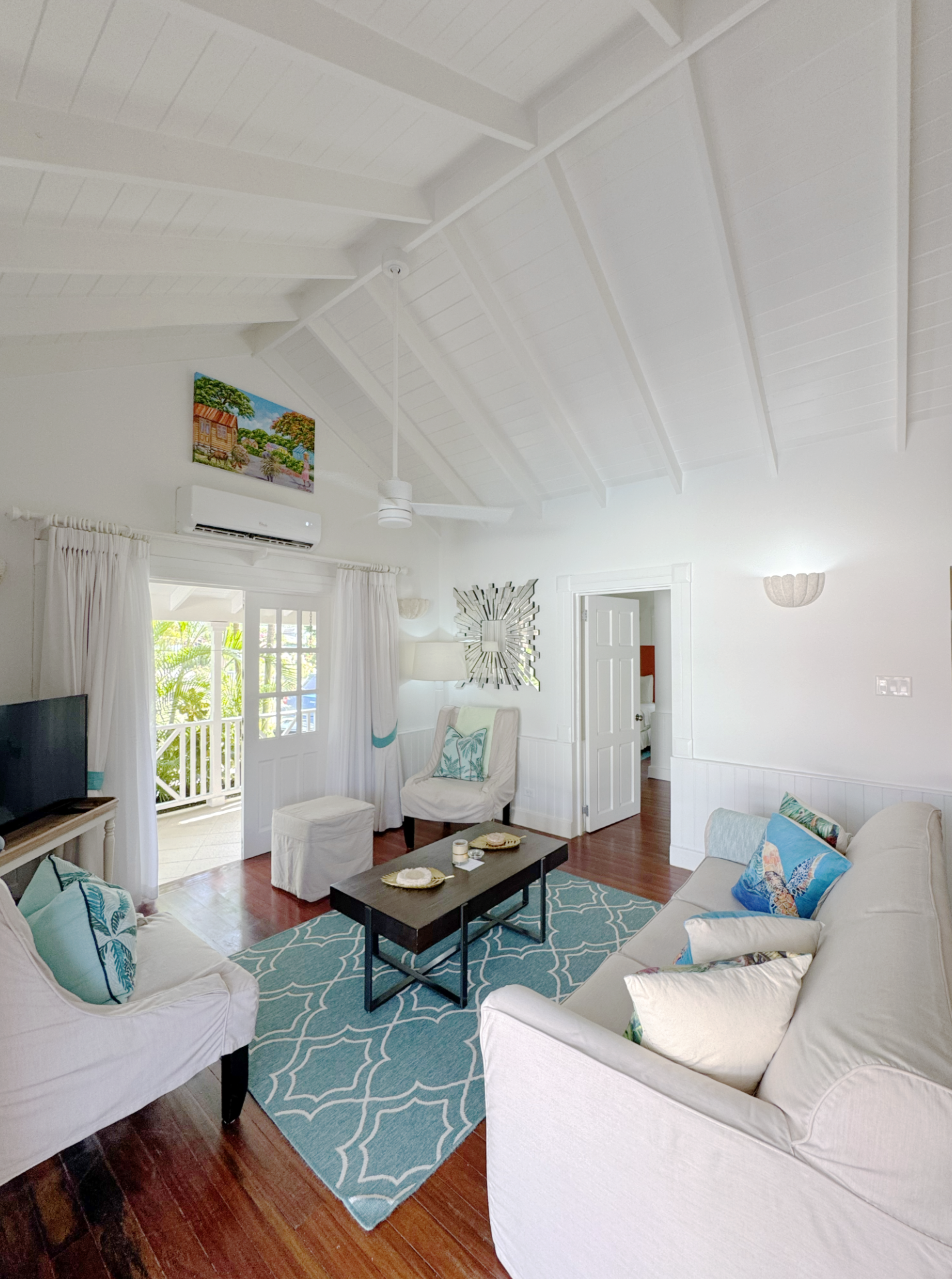 Living room with white wicker sofas, a turquoise patterned rug, a dark coffee table, and a ceiling fan. There are pillows with tropical motifs on the sofas. Large windows with white curtains let in natural light. The room has white walls and ceiling,