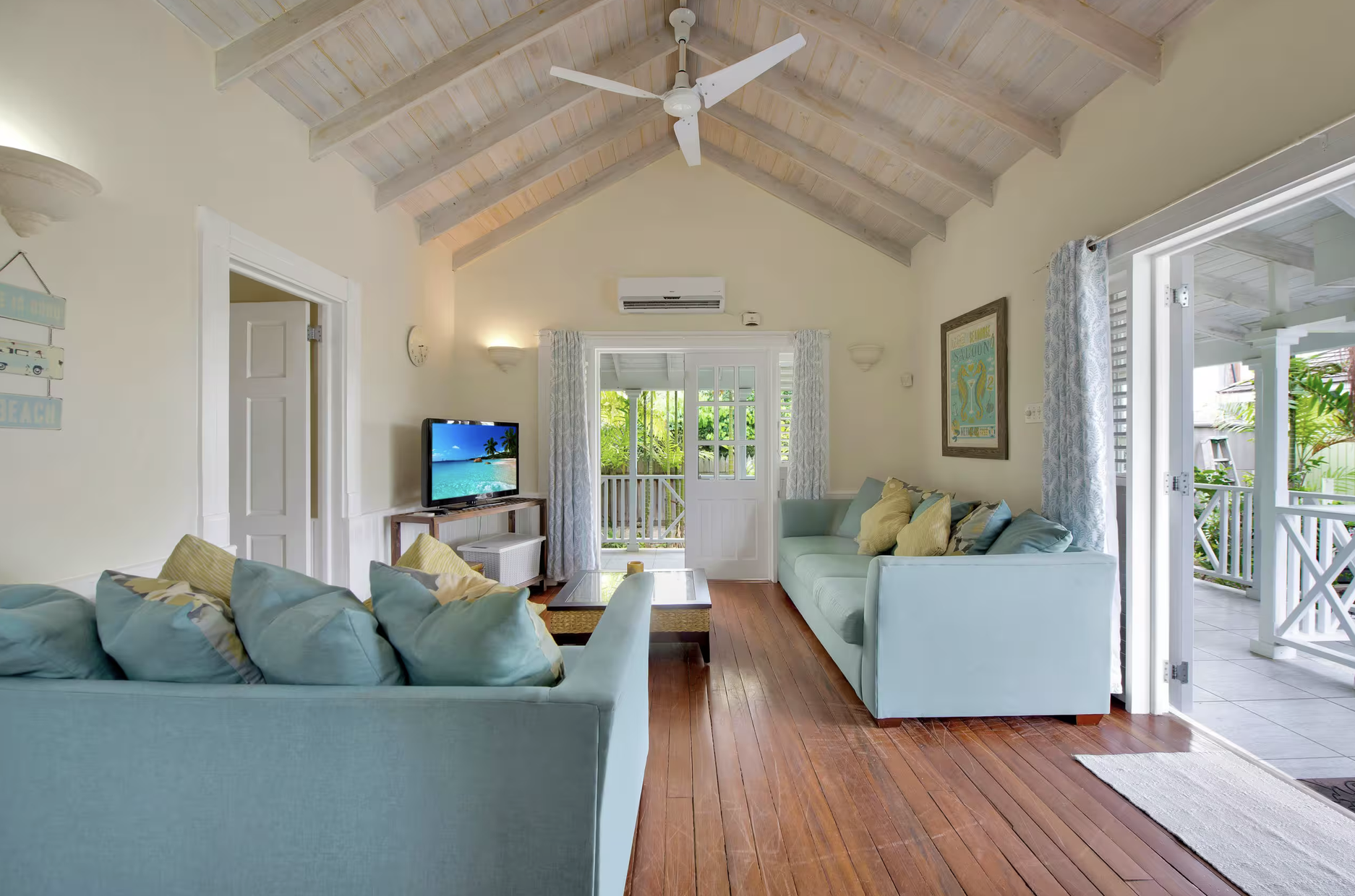 Living room with light-colored sofas, a wooden coffee table, a small TV showing a beach scene, and sliding glass doors leading to an outdoor balcony with greenery.