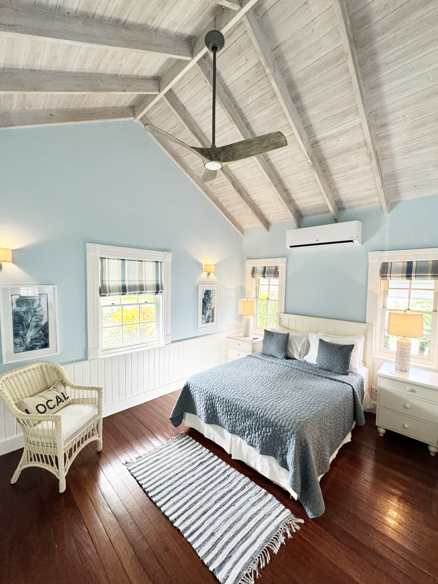 A bedroom with light blue walls, a wooden vaulted ceiling, and wooden floors. There is a bed with a blue quilt and white bed skirt, with two blue pillows and white pillows. Two nightstands with lamps and artwork are on either side of the bed. There a