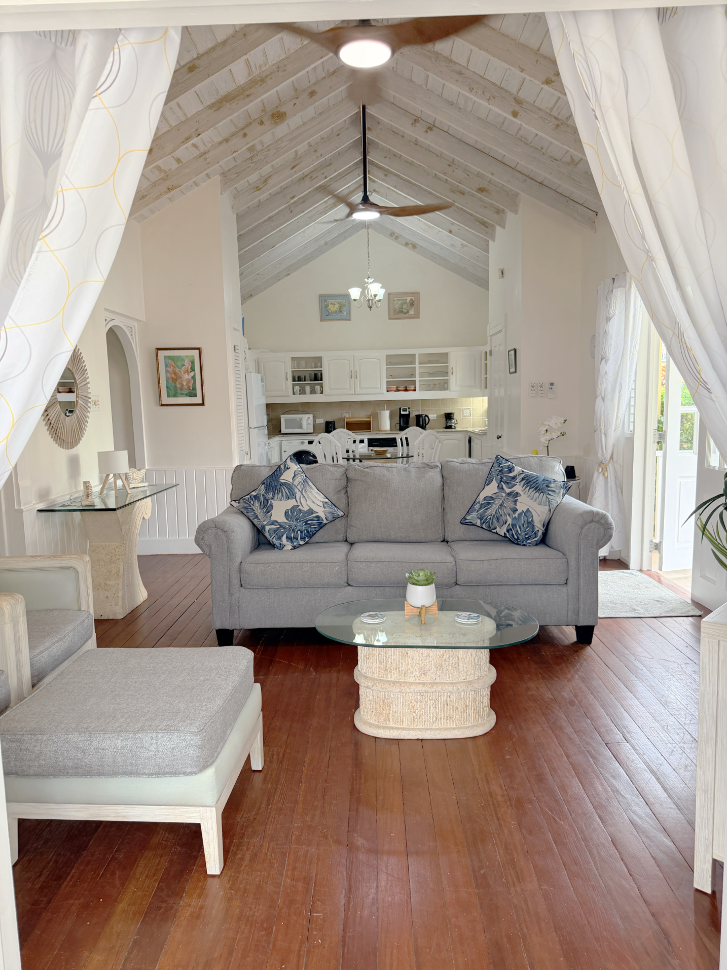 A cozy living room with a gray sofa, patterned pillows, a glass coffee table, and wooden flooring. The background shows a kitchen with white cabinets, a microwave, and decorative wall art. Curtains frame a doorway to the outside, and a ceiling fan an