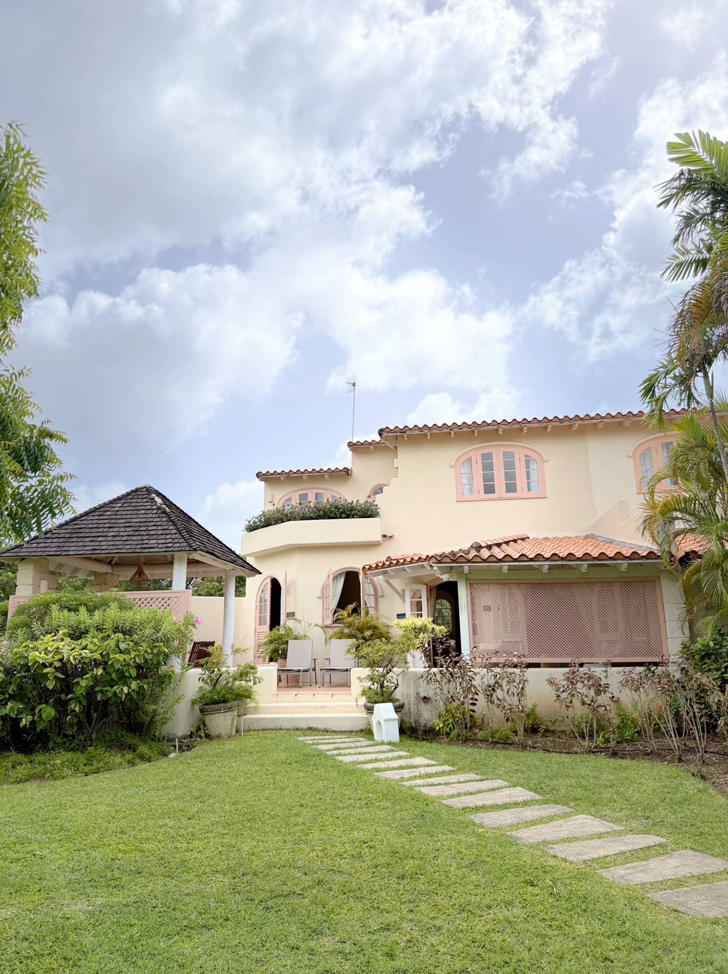 A house with a pink and white exterior, surrounded by lush green plants and a manicured lawn. The house has arched windows and a tiled roof, with a patio area and outdoor seating.