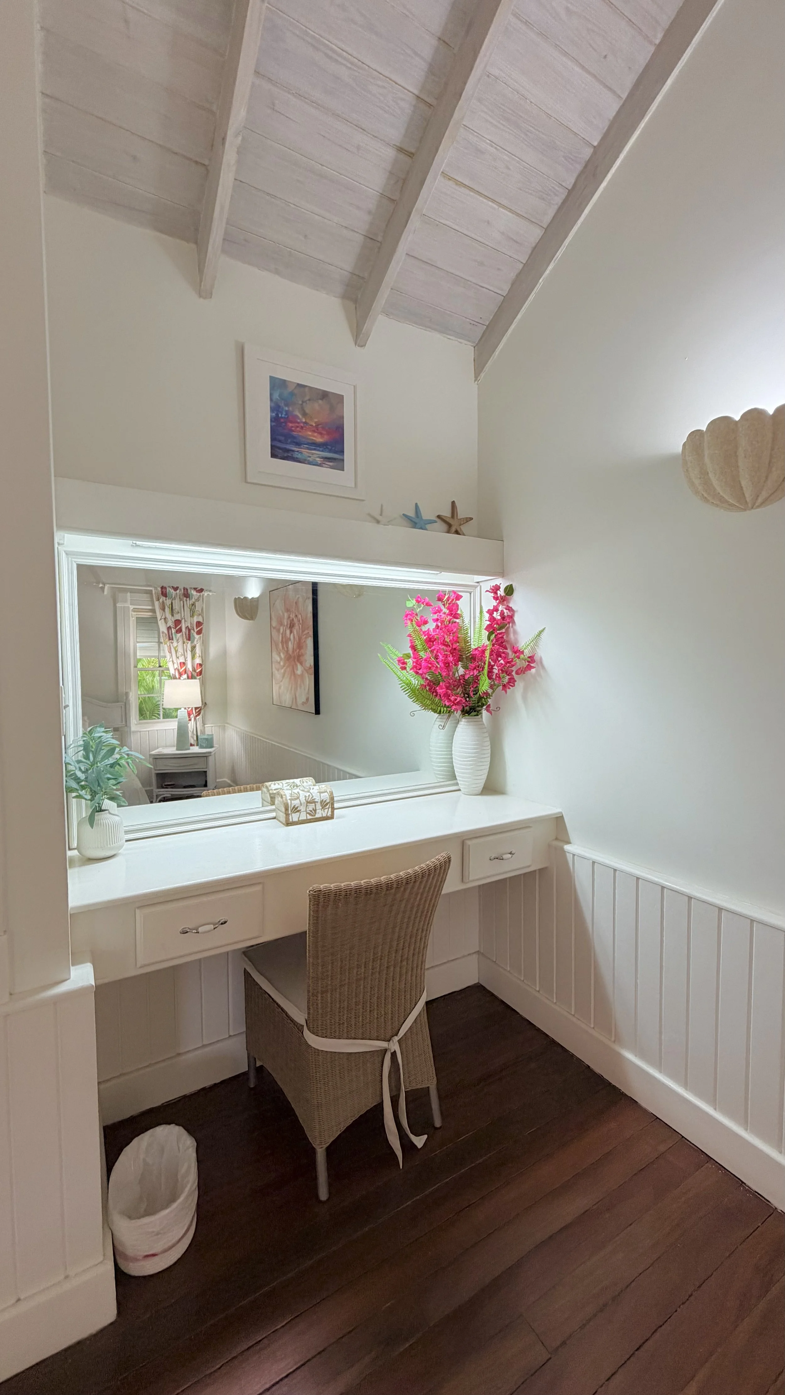 A cozy corner with a white vanity table and matching chair, decorated with a large vase of pink flowers, a smaller vase with green leaves, and a decorative box. A large mirror is mounted on the wall above, reflecting a room with a window, curtains, a