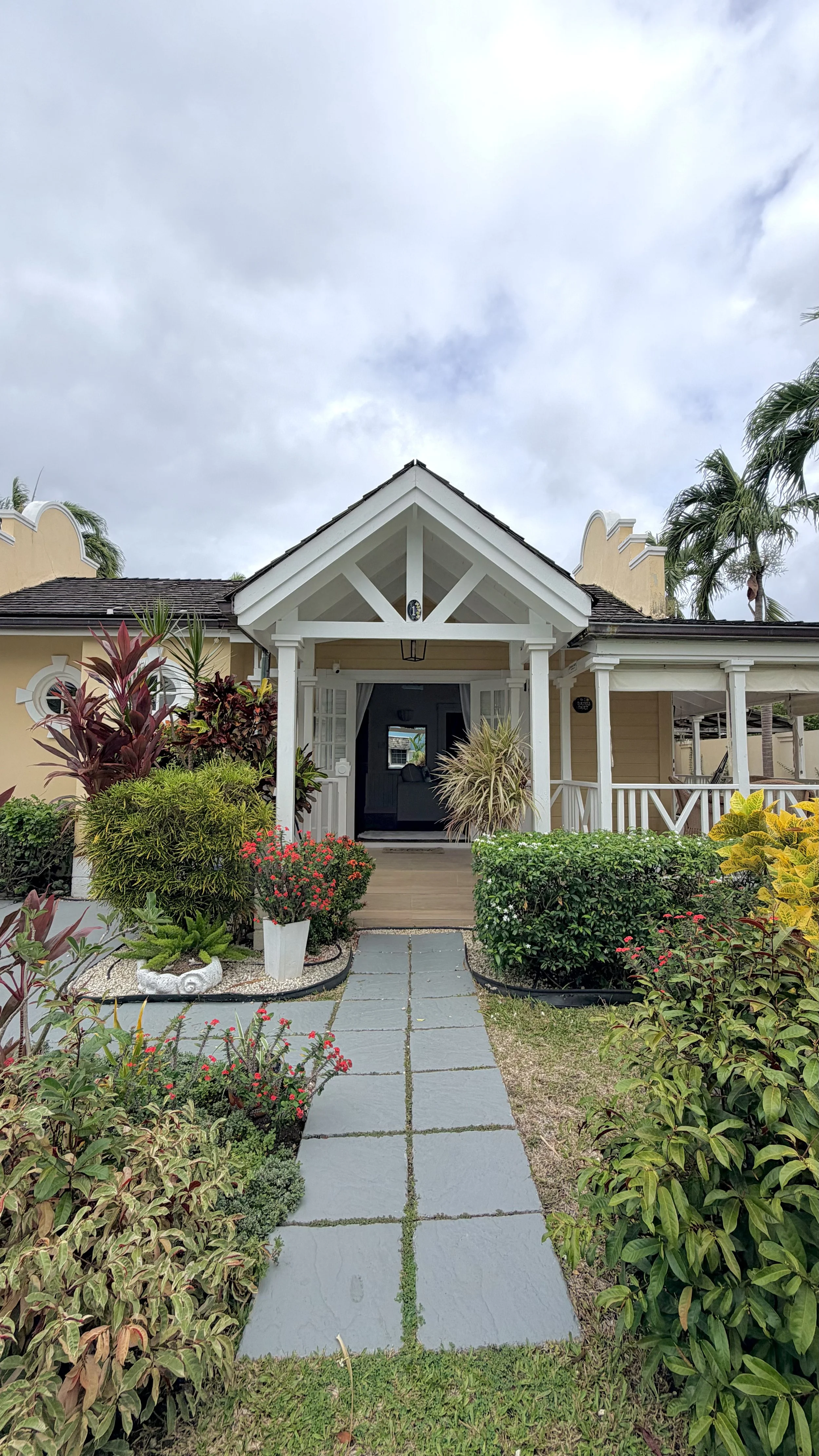 Front view of a yellow house with a porch, surrounded by lush green plants and colorful flowers, with a stone pathway leading to the entrance and palm trees in the background under a cloudy sky.
