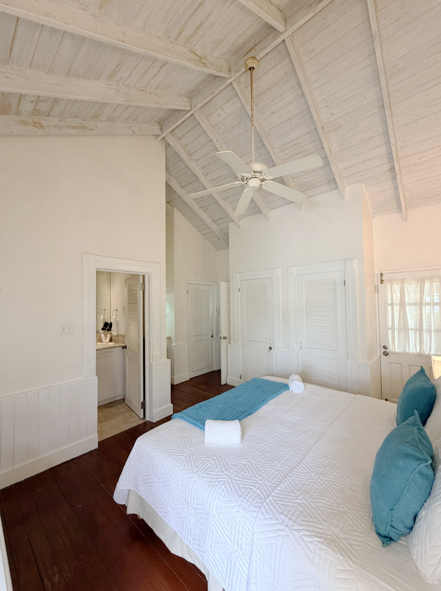 A bedroom with a white bed, blue pillows, and a rolled white towel. The room has white walls, louvered closet doors, and a high, white wooden ceiling with a ceiling fan.