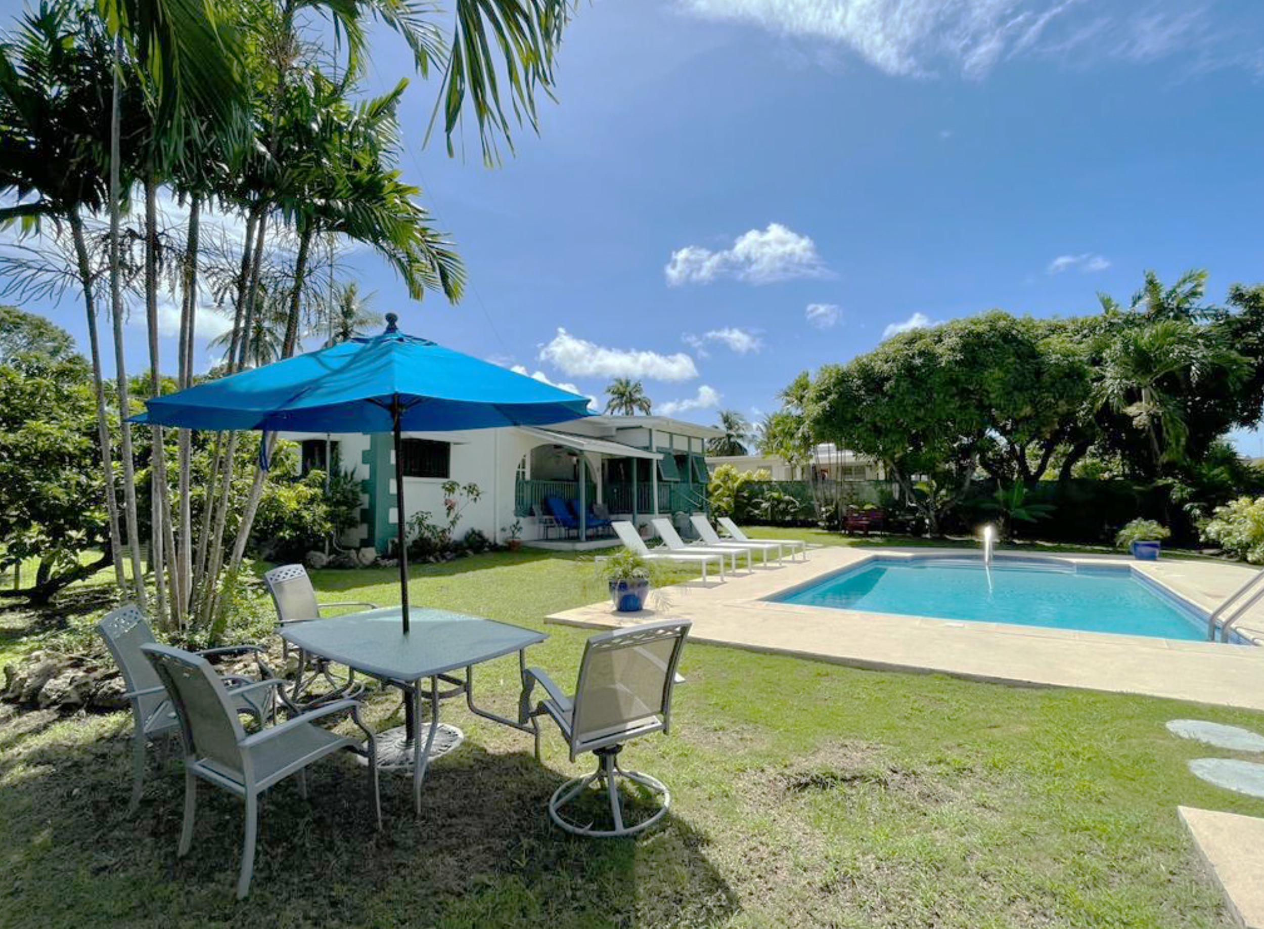 A backyard with a swimming pool, surrounded by green grass and trees, with patio furniture and umbrellas, and a bright sky with some clouds.