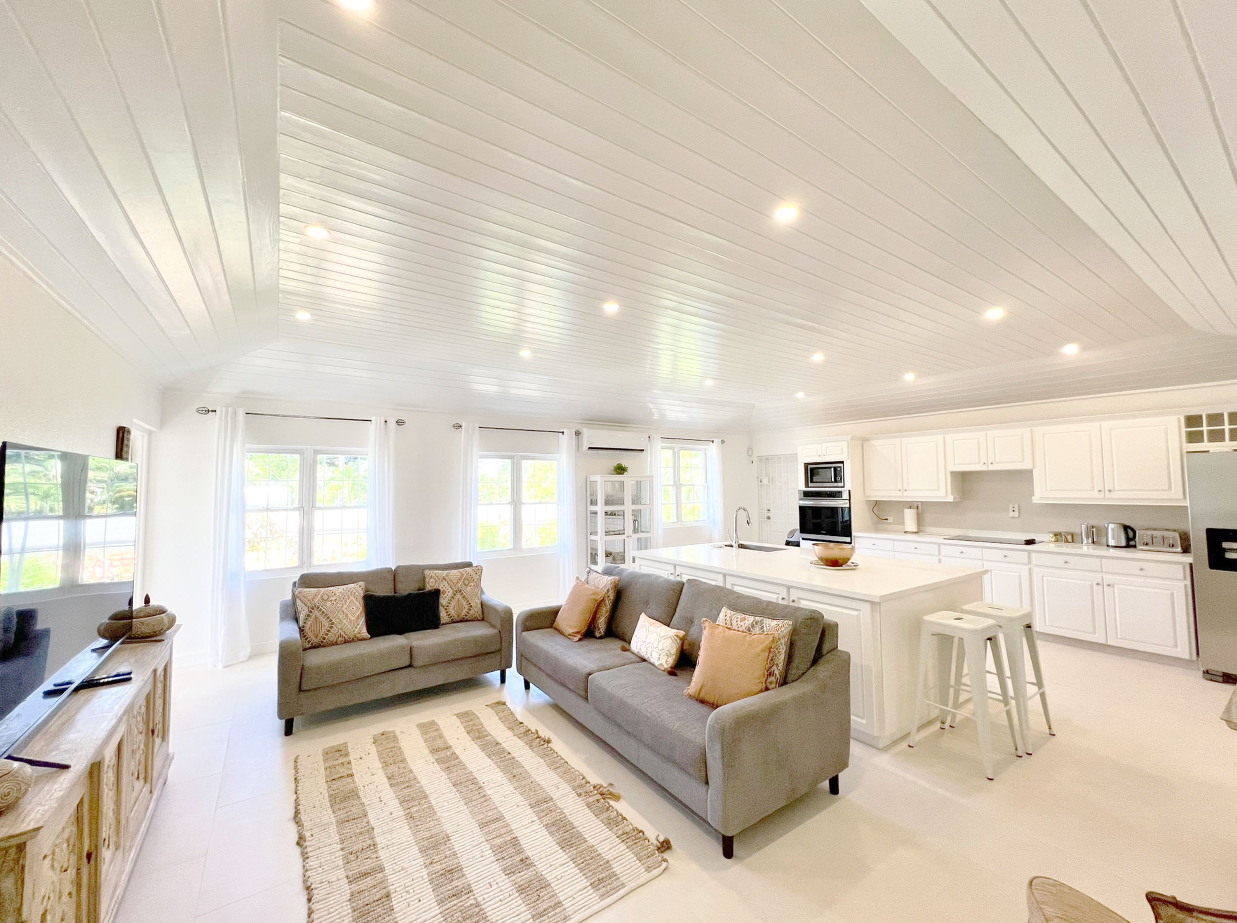 Living room with white ceiling, recessed lighting, large windows with white curtains, a brown sofa with patterned pillows, a wooden TV stand, a rug, and part of a kitchen with white cabinets and a countertop.
