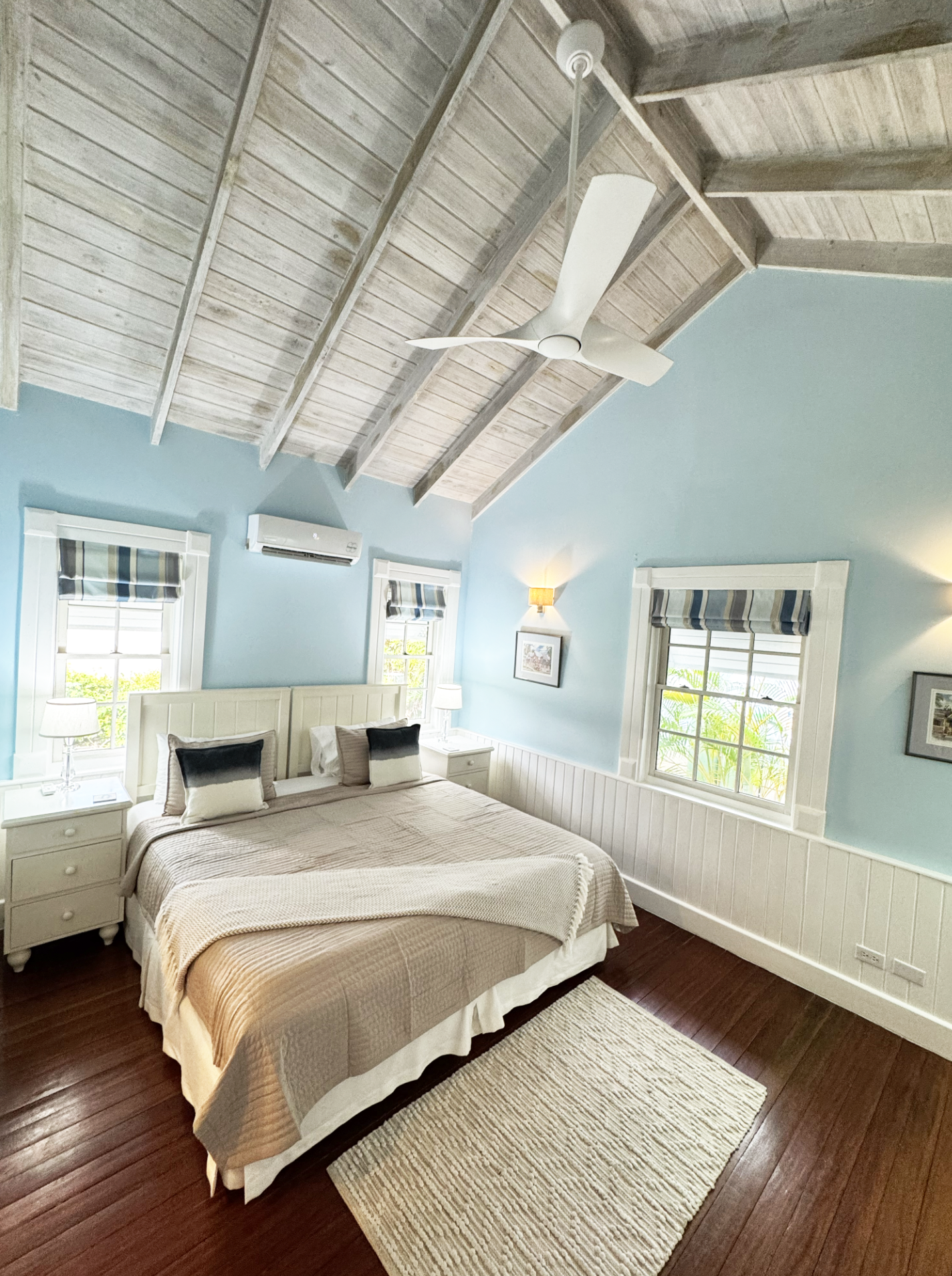 A bedroom with a high, wooden, vaulted ceiling with a modern white ceiling fan, two windows with plaid blinds, a beige bed with white and black pillows, white nightstands with lamps, a framed picture on the wall, and a beige area rug on dark hardwood