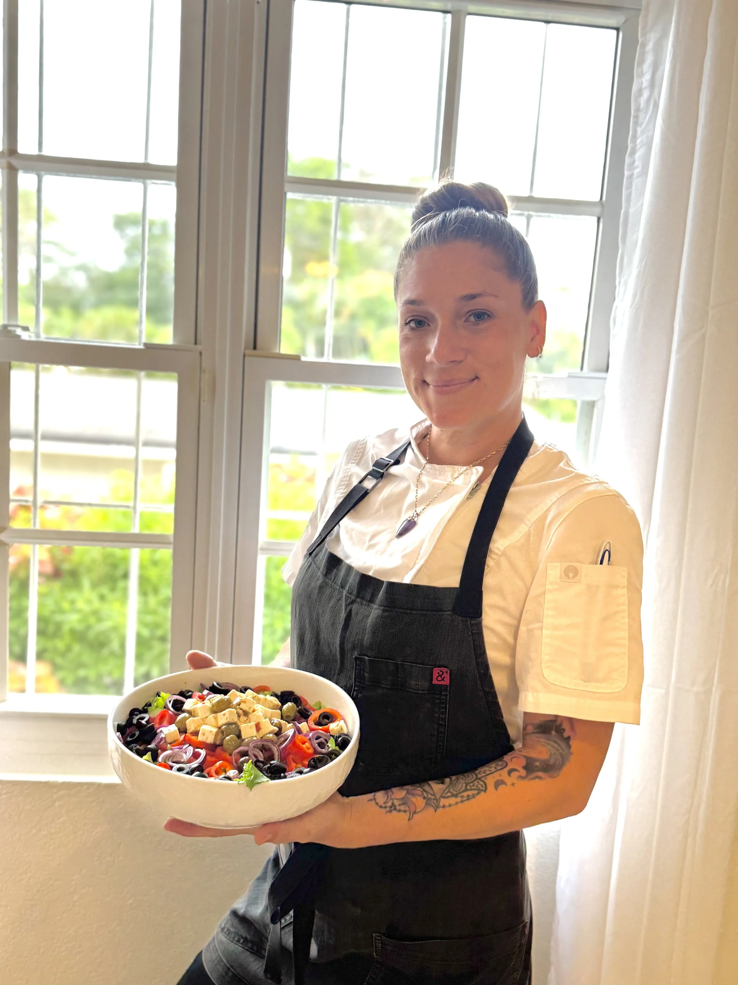 A woman with tattoos on her arm wearing a yellow shirt, black apron, holding a large bowl of colorful pasta salad, standing by a window with greenery outside.